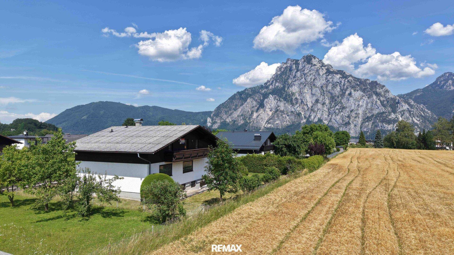 Panoramablick auf ein Einfamilienhaus mit Garten, umgeben von Feldern und majestätischen Bergen.