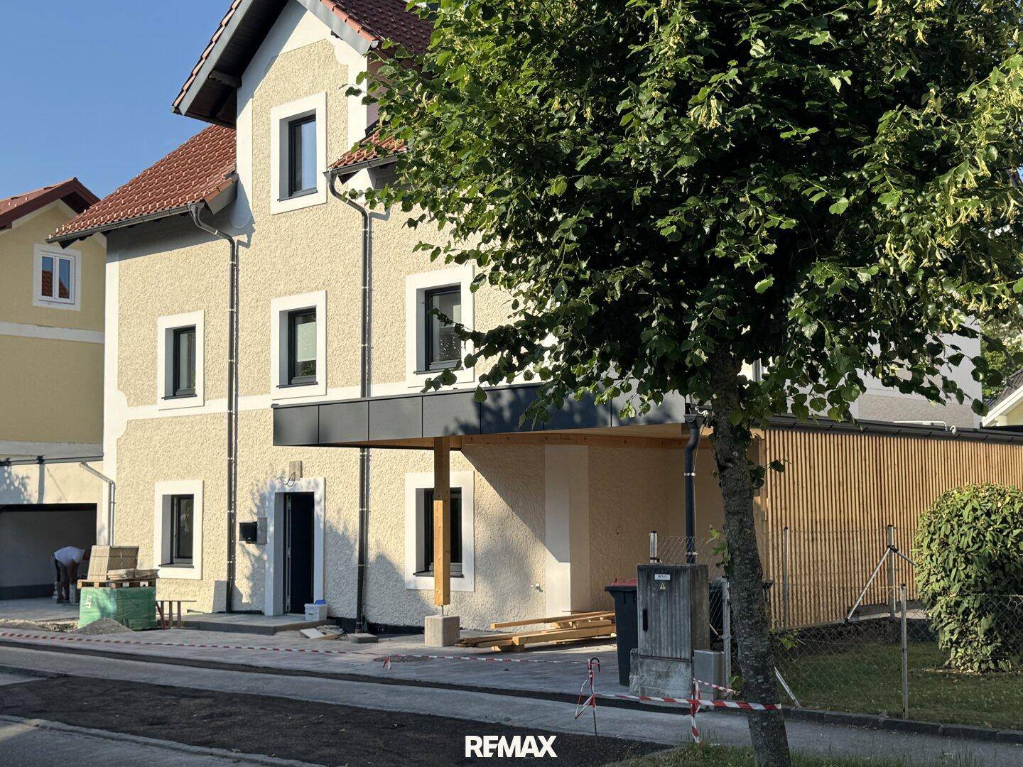 Seitenansicht eines Neubaus mit Carport und moderner Holzverkleidung unter einem Baum.