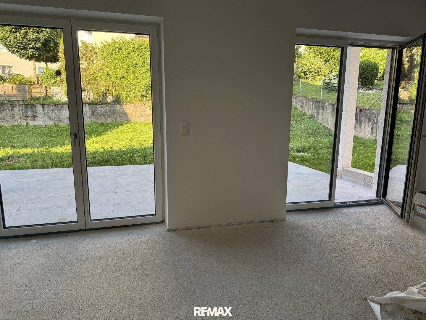 Unfertiger Innenraum mit großen Fensterfronten und direktem Zugang zur Terrasse und Garten.