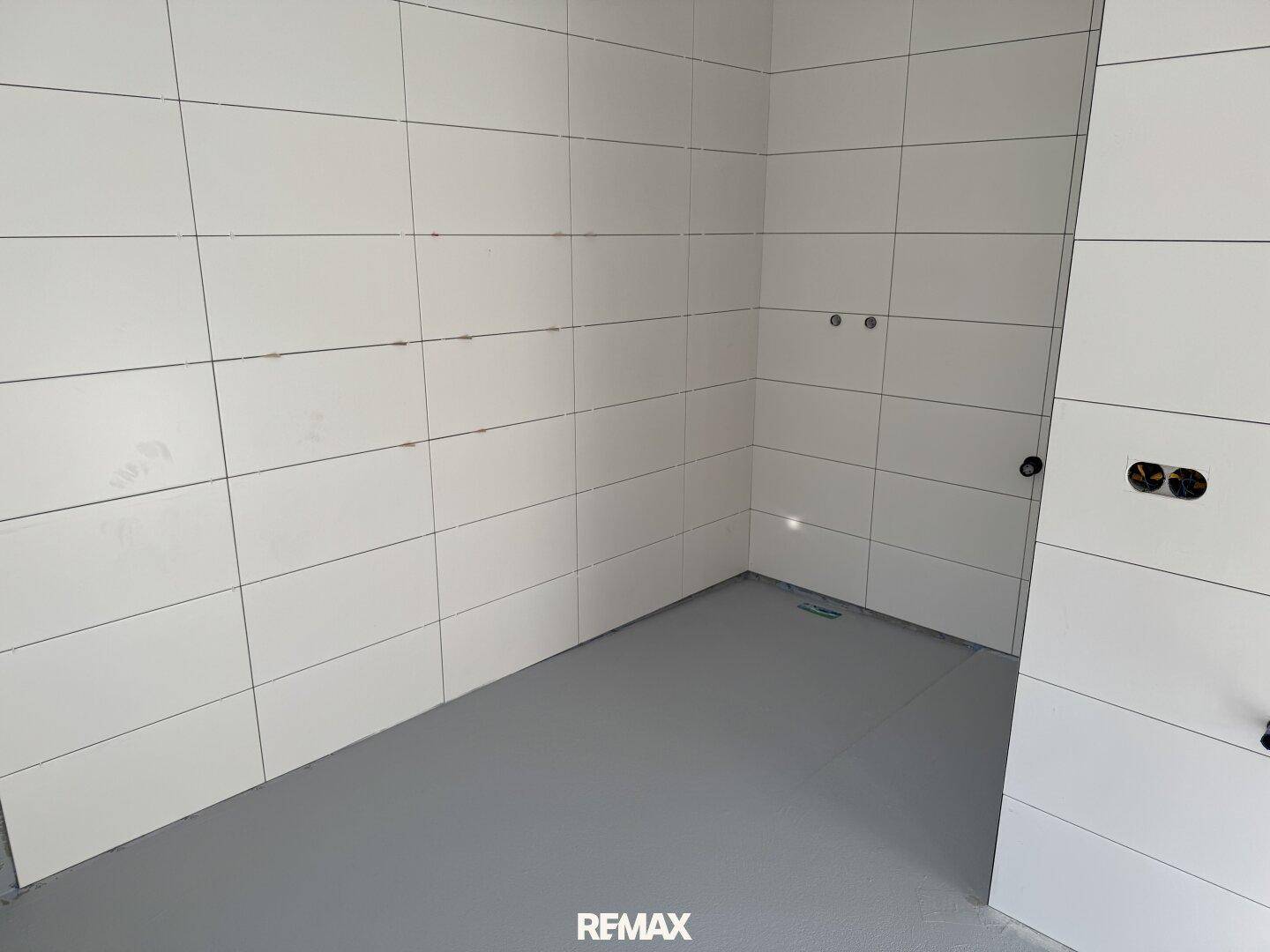 Unfertiges Badezimmer mit weißen Fliesen an den Wänden und vorbereiteten Anschlüssen für Sanitäranlagen.