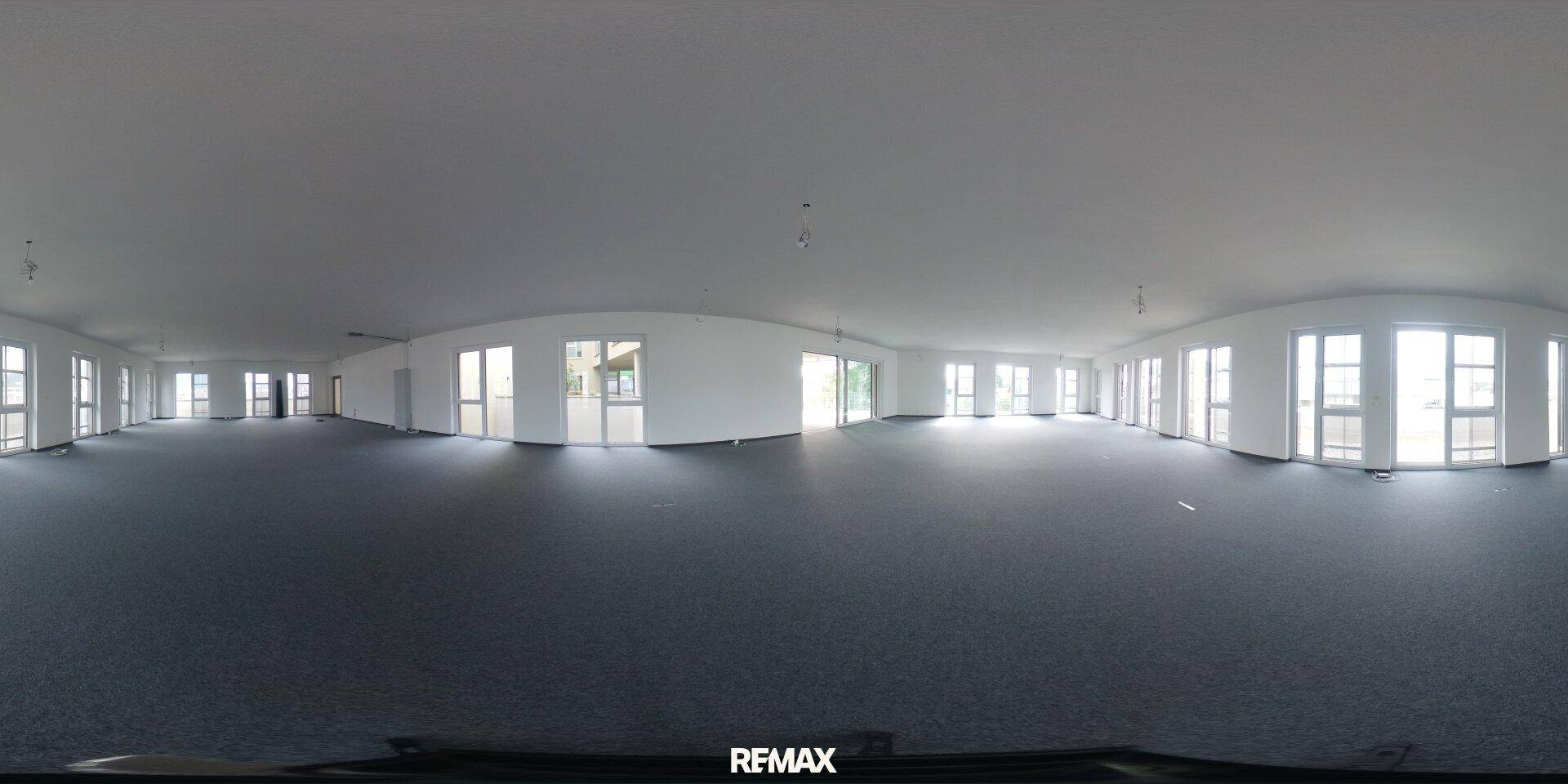 Panoramaansicht eines weitläufigen, leeren Büroraums mit grauem Teppichboden und vielen Fenstern.