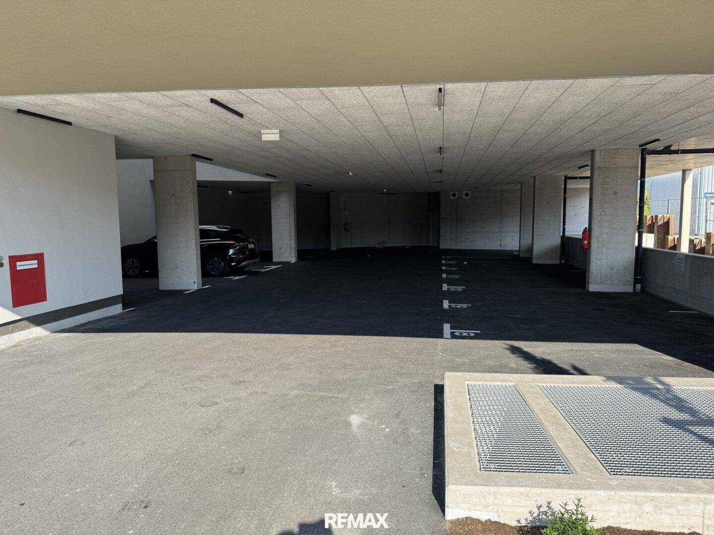 Überdachter Parkbereich mit Betonstützen und Zufahrt zu weiteren Stellplätzen, Teil eines modernen Gebäudekomplexes.
