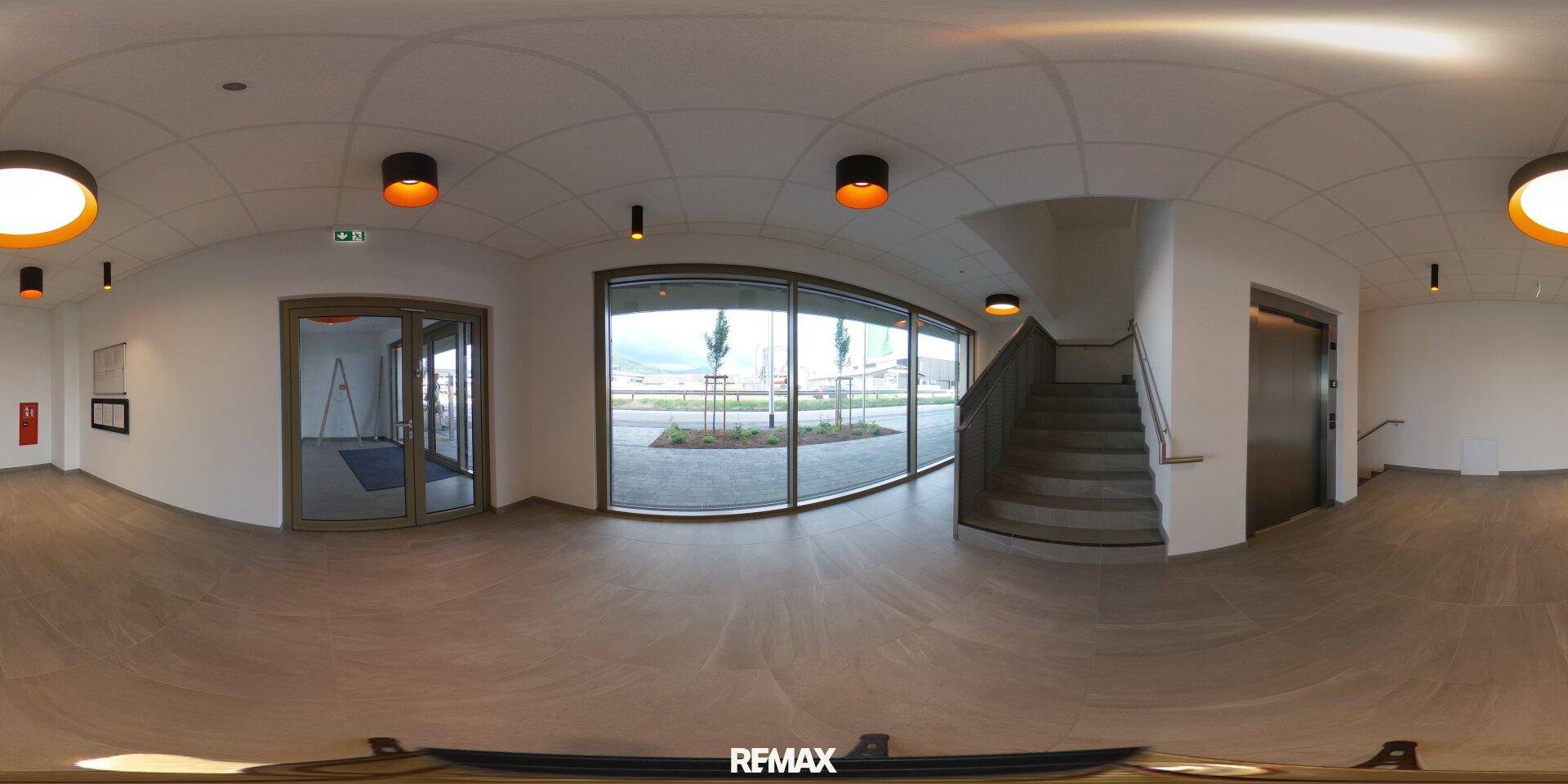Panoramaansicht eines modernen Eingangsbereichs mit großen Glasfronten, Treppe und Aufzug.