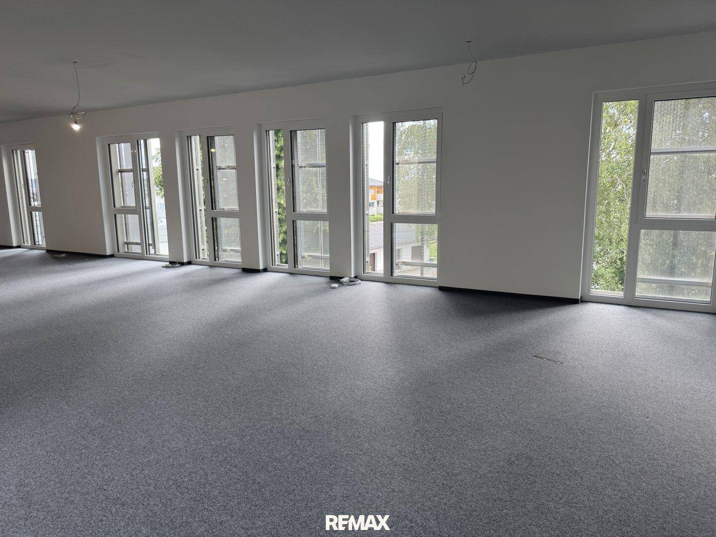Langer Büroraum mit vielen Fenstern und dunklem Vinylboden, ideal für Großraumbüros.