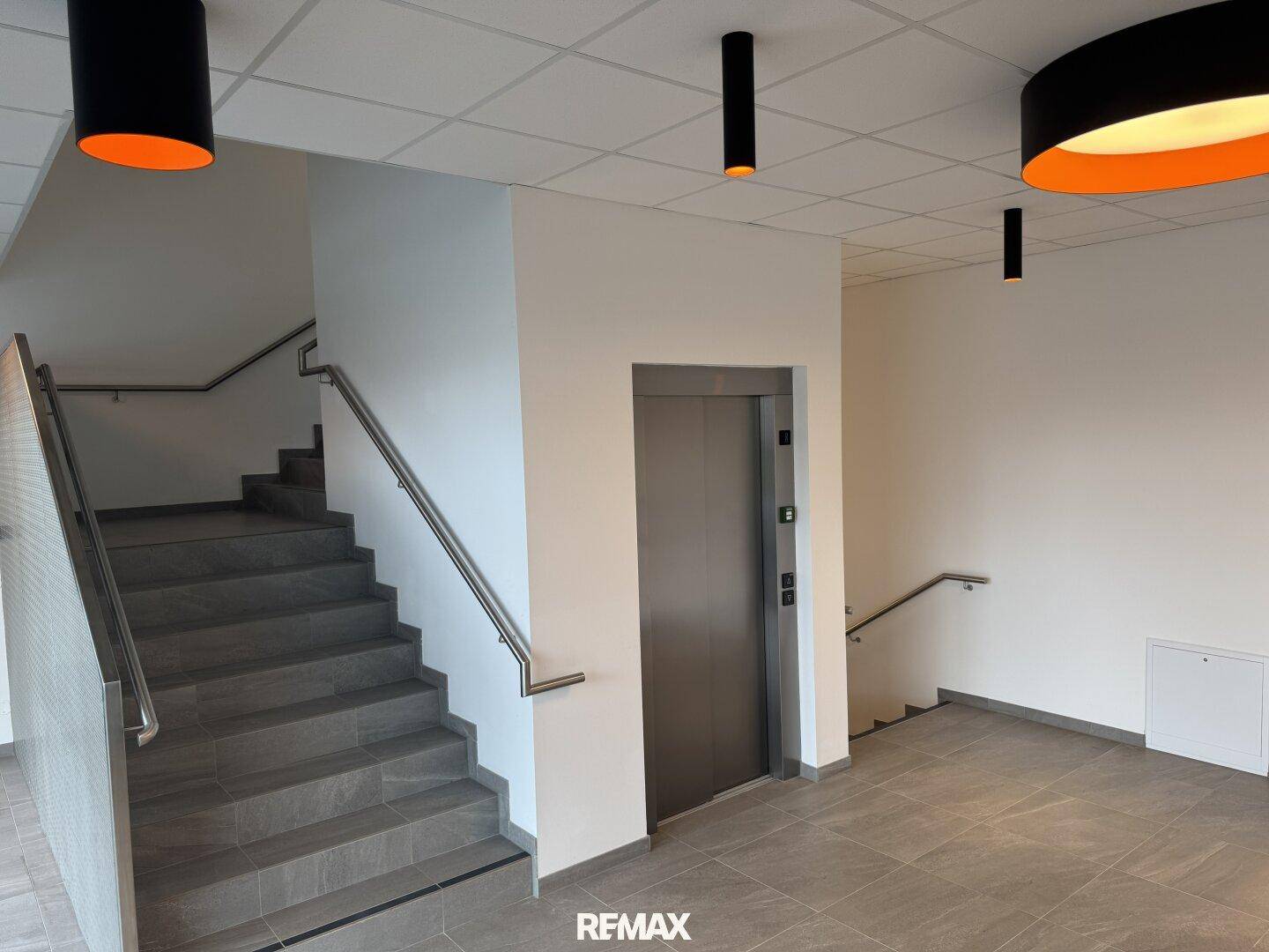 Heller Eingangsbereich mit Treppe, Aufzug und modernen Deckenleuchten.