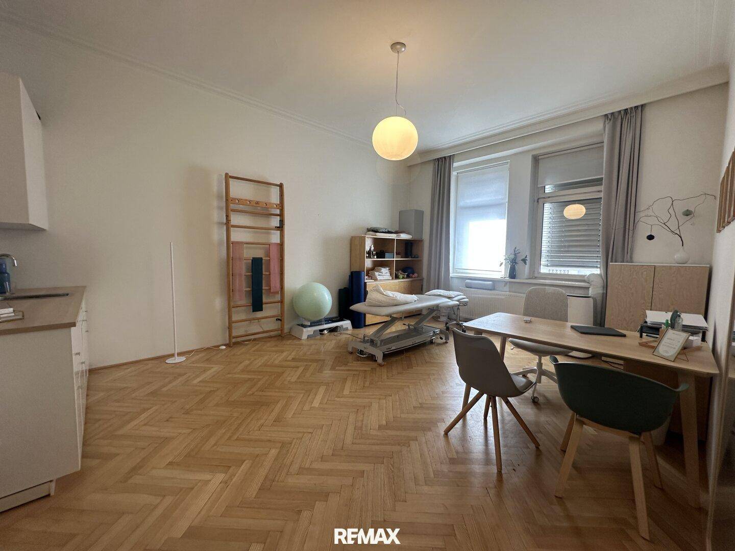 Geräumiges Büro oder Therapiezimmer mit Fischgrätparkett, Schreibtisch und Behandlungsbank unter hohen Decken.