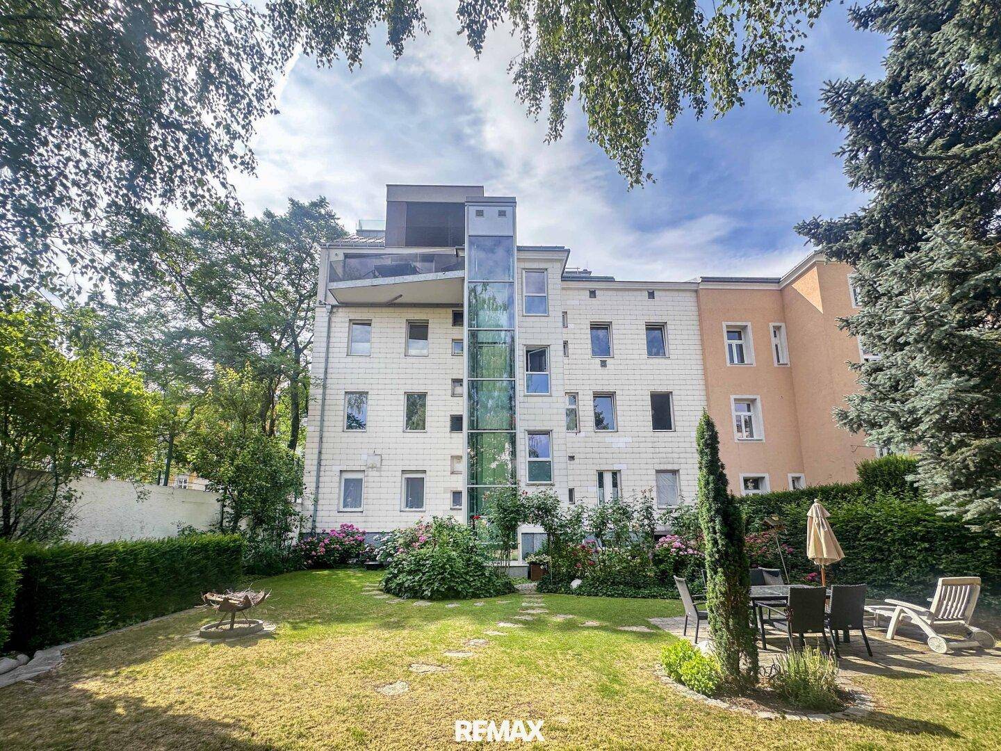 Großer gepflegter Garten mit Rasenfläche, Sitzbereich und einem externen Glasaufzug am modernen Gebäude.