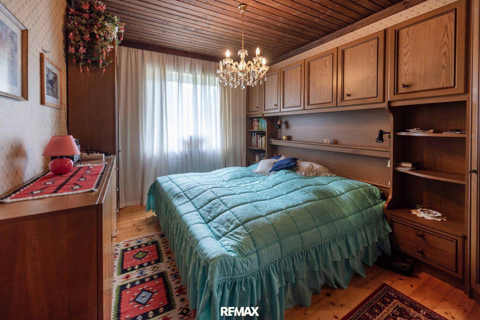 Schlafzimmer mit Holzdecke, Einbauschränken und einem großen Bett mit türkiser Tagesdecke.