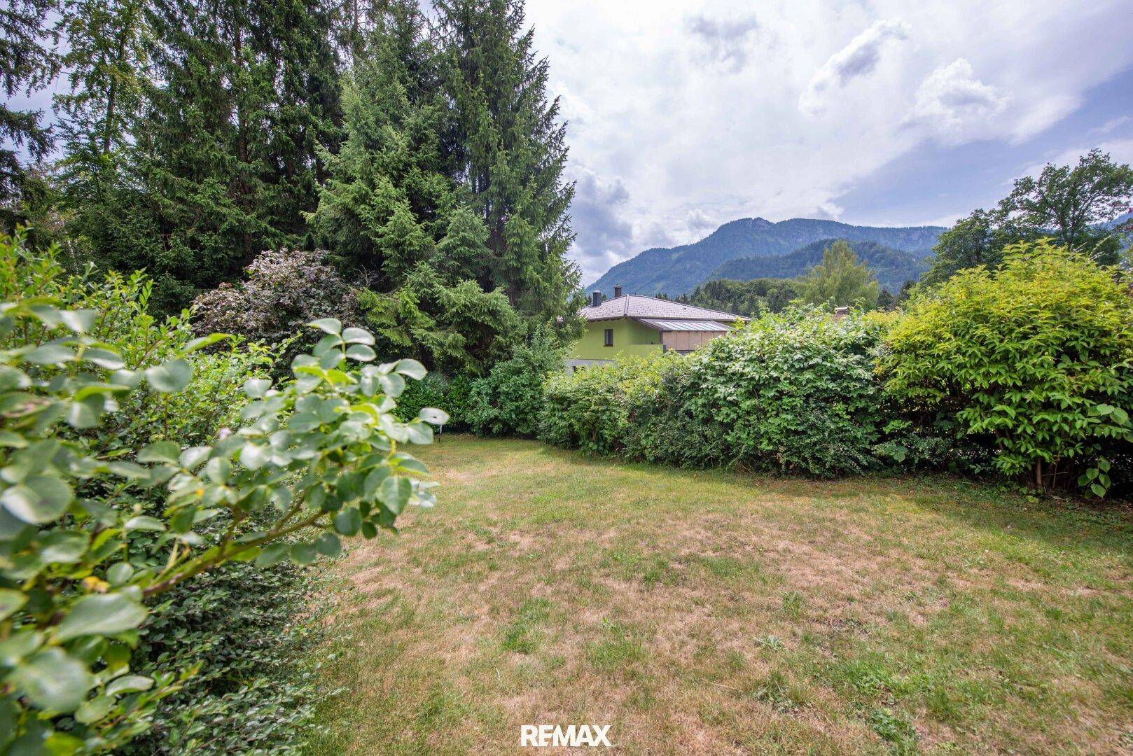 Großer Gartenbereich mit Rasenfläche, umgeben von Bäumen und Sträuchern mit Bergblick.