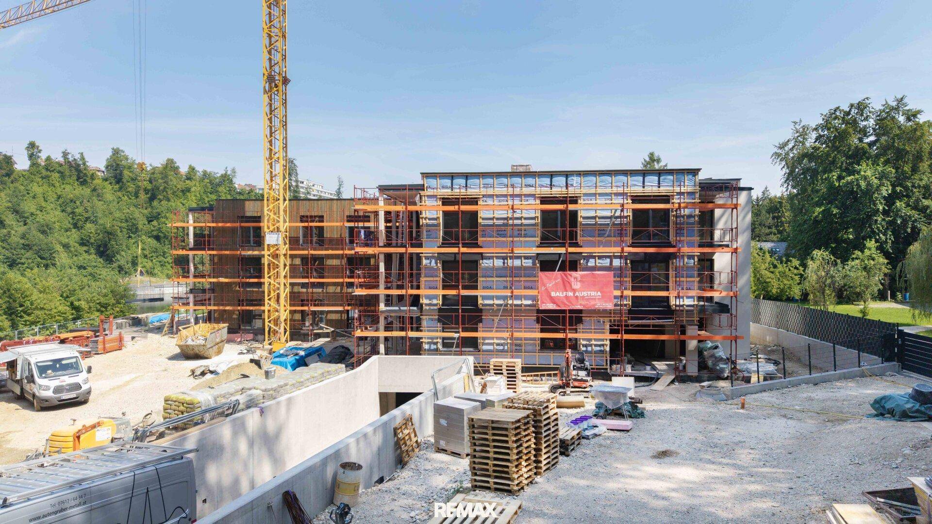 Baustelle eines Neubaus mit Kran, Tiefgaragenzufahrt und umliegender Natur im Sonnenlicht.