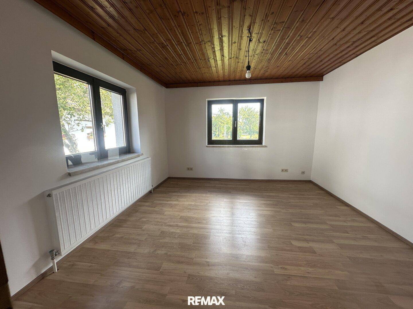 Heller Raum mit Holzdeckenverkleidung, Laminatboden und zwei Fenstern.