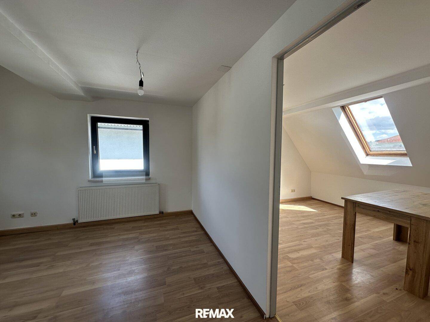 Heller Raum mit Laminatboden, Dachschräge und Blick auf ein Dachfenster im Nebenraum.