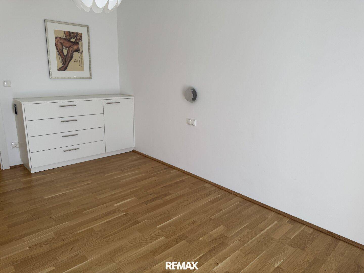 Helles Zimmer mit Holzparkettboden und einer weißen Kommode an der Wand.