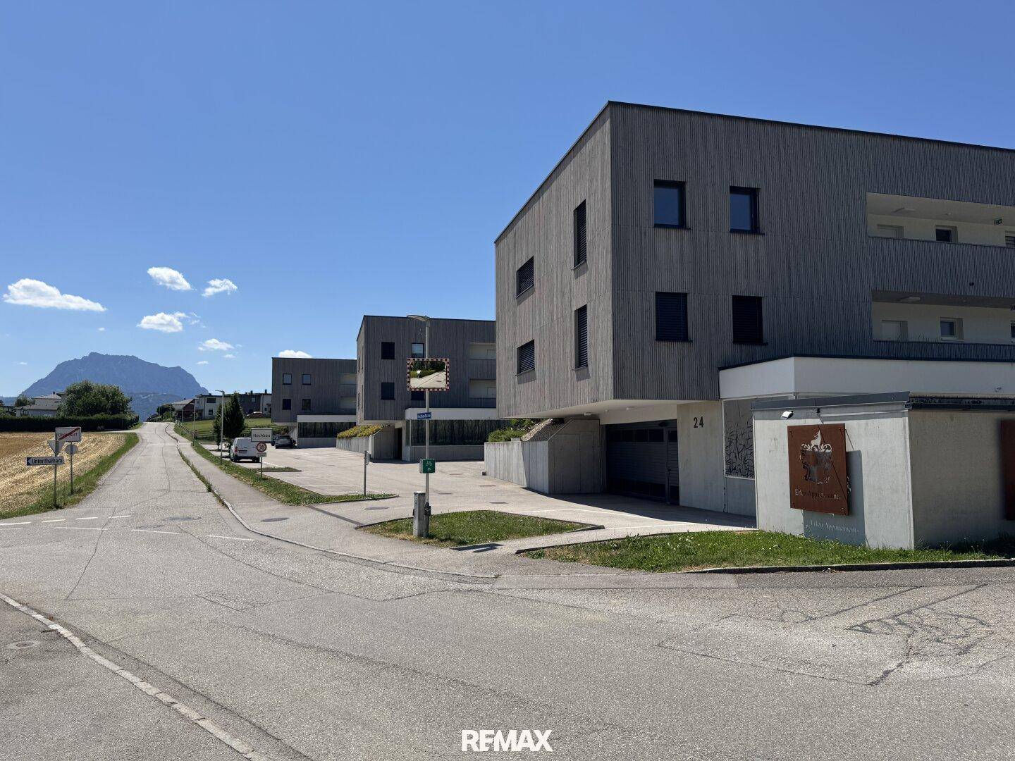 Straße mit modernen Gebäuden, Feldern und einem Berg im Hintergrund unter blauem Himmel.