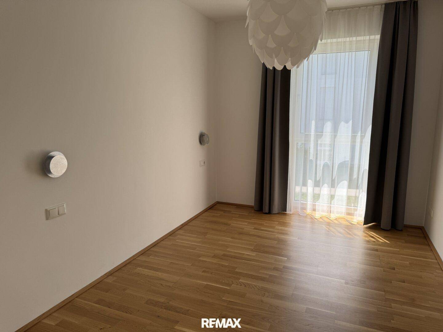 Lichtdurchflutetes Zimmer mit Holzparkett, großen Fenstern und moderner Deckenleuchte.