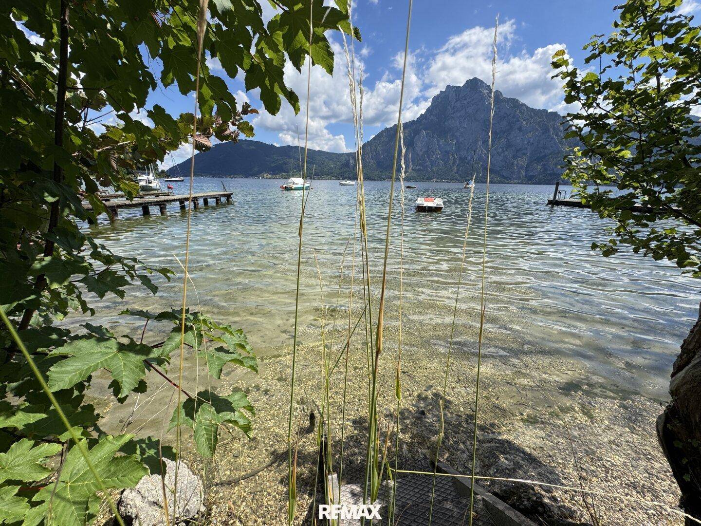 Klares Seewasser mit sichtbarem Kiesgrund, umrahmt von grünen Blättern und mit Blick auf die Berge.
