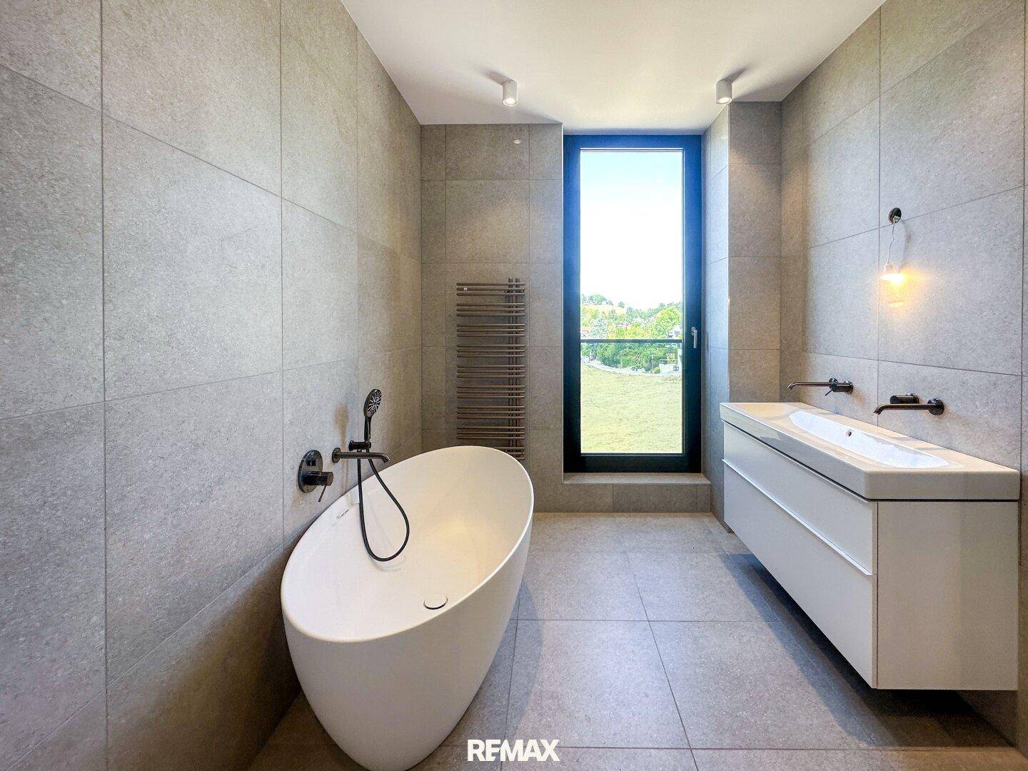 Modernes Badezimmer mit freistehender Badewanne, Doppelwaschbecken und großem Fenster mit Grünblick.