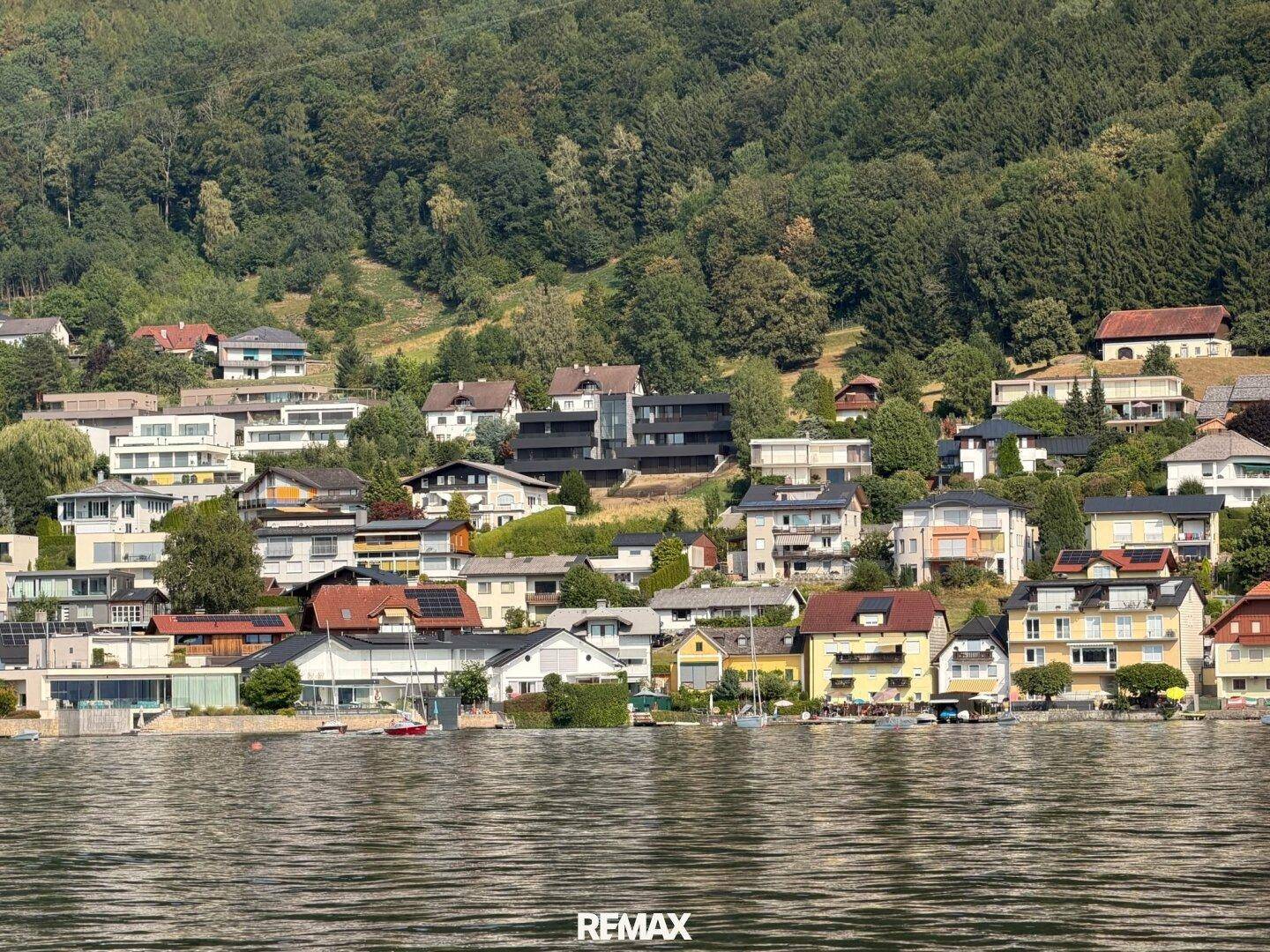 Malerische Ansicht der Häuser am Seeufer mit bewaldeten Hügeln im Hintergrund, typisch für die Region.