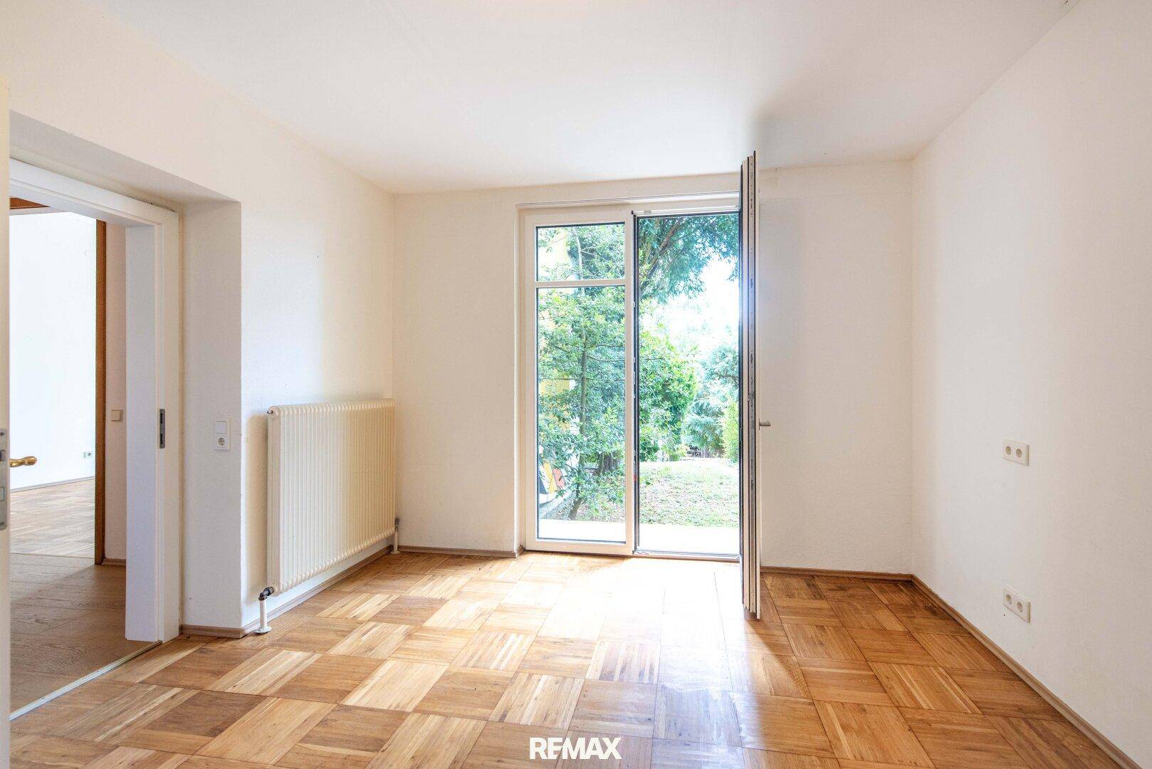 Helles Zimmer mit Parkettboden und direktem Zugang zu einem grünen Außenbereich.