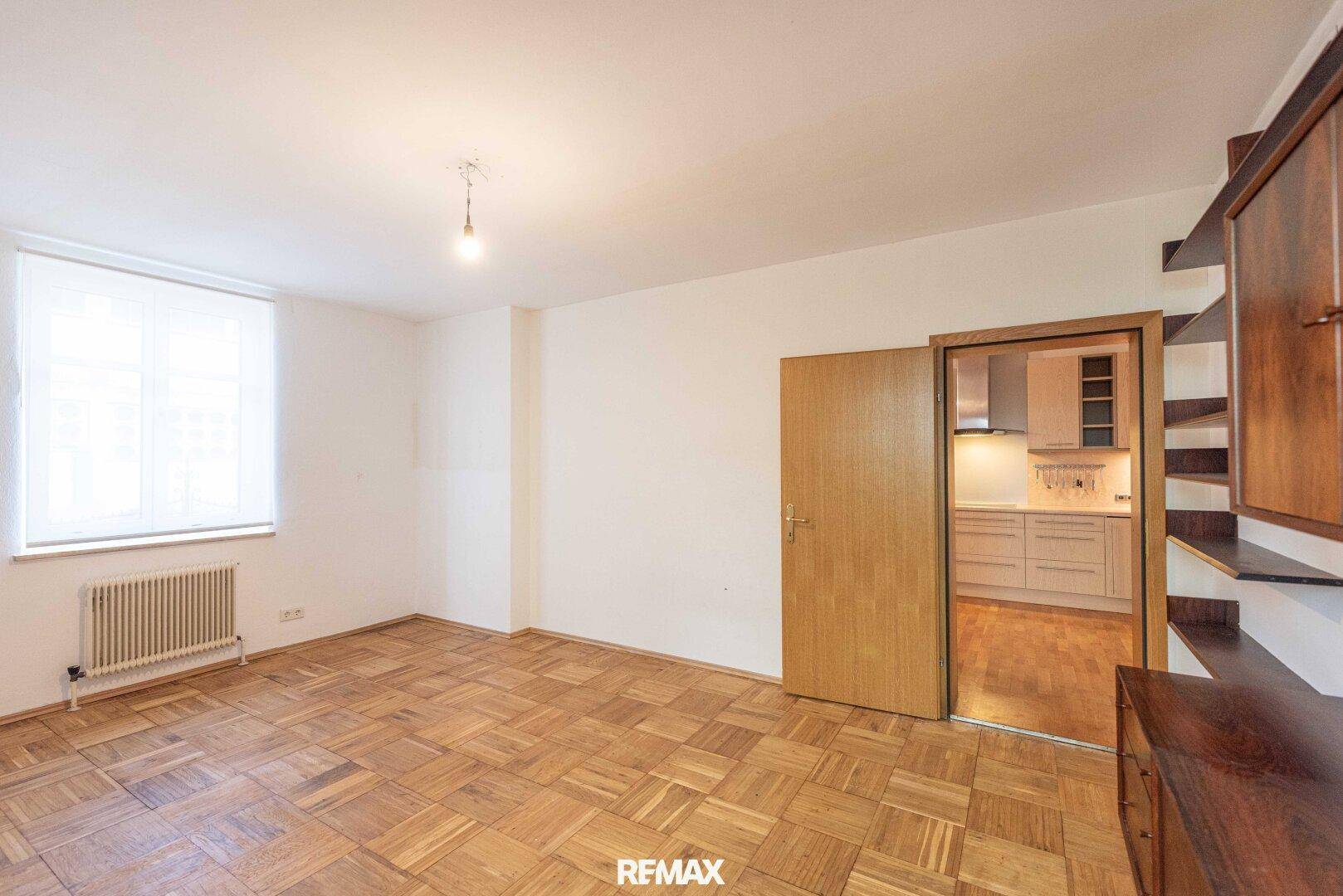 Geräumiges Zimmer mit Parkettboden und Fenster, das zur Küche hin offen ist.