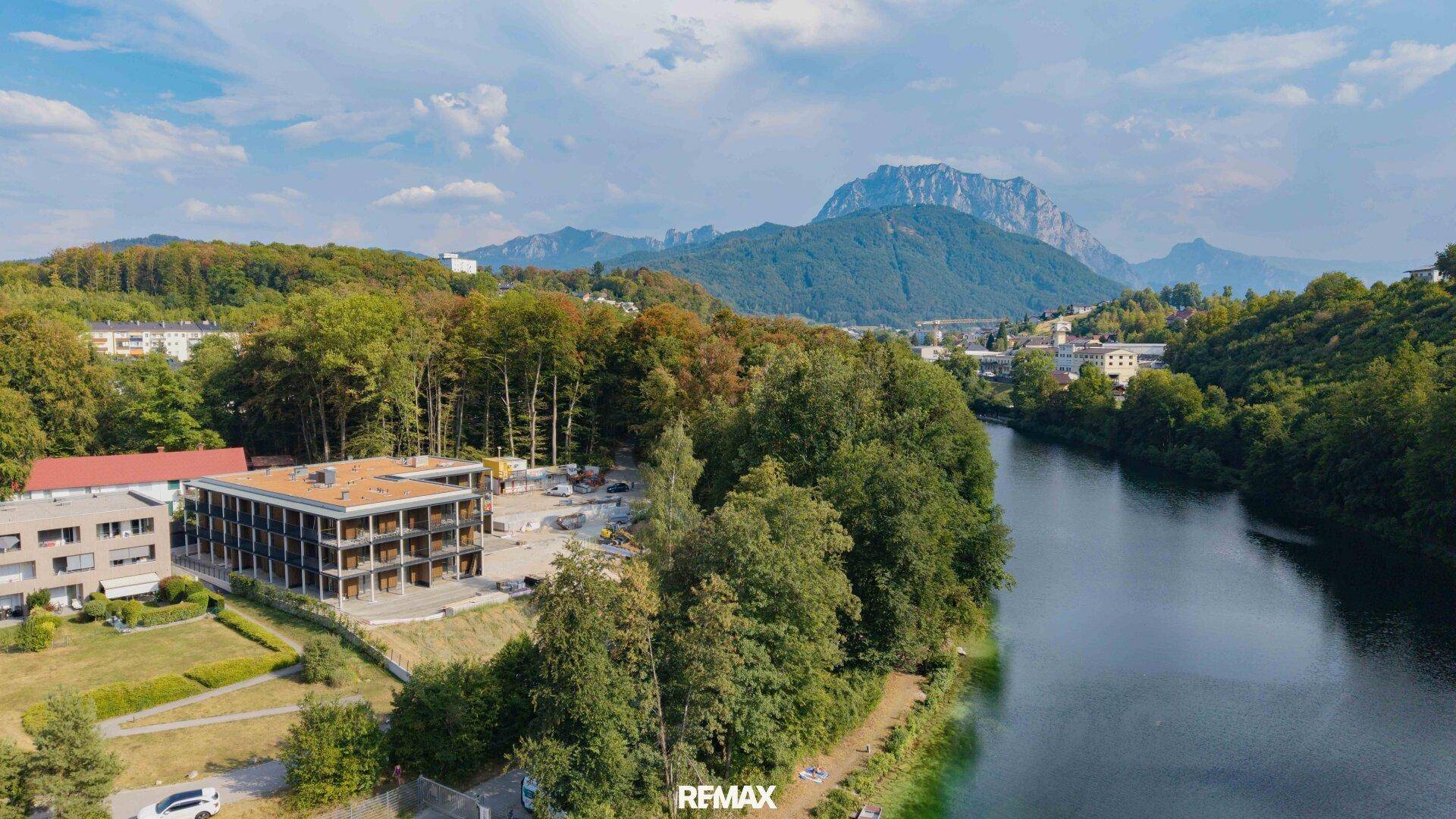 Luftaufnahme des modernen Gebäudes am Flussufer mit Bergblick und viel Grün.