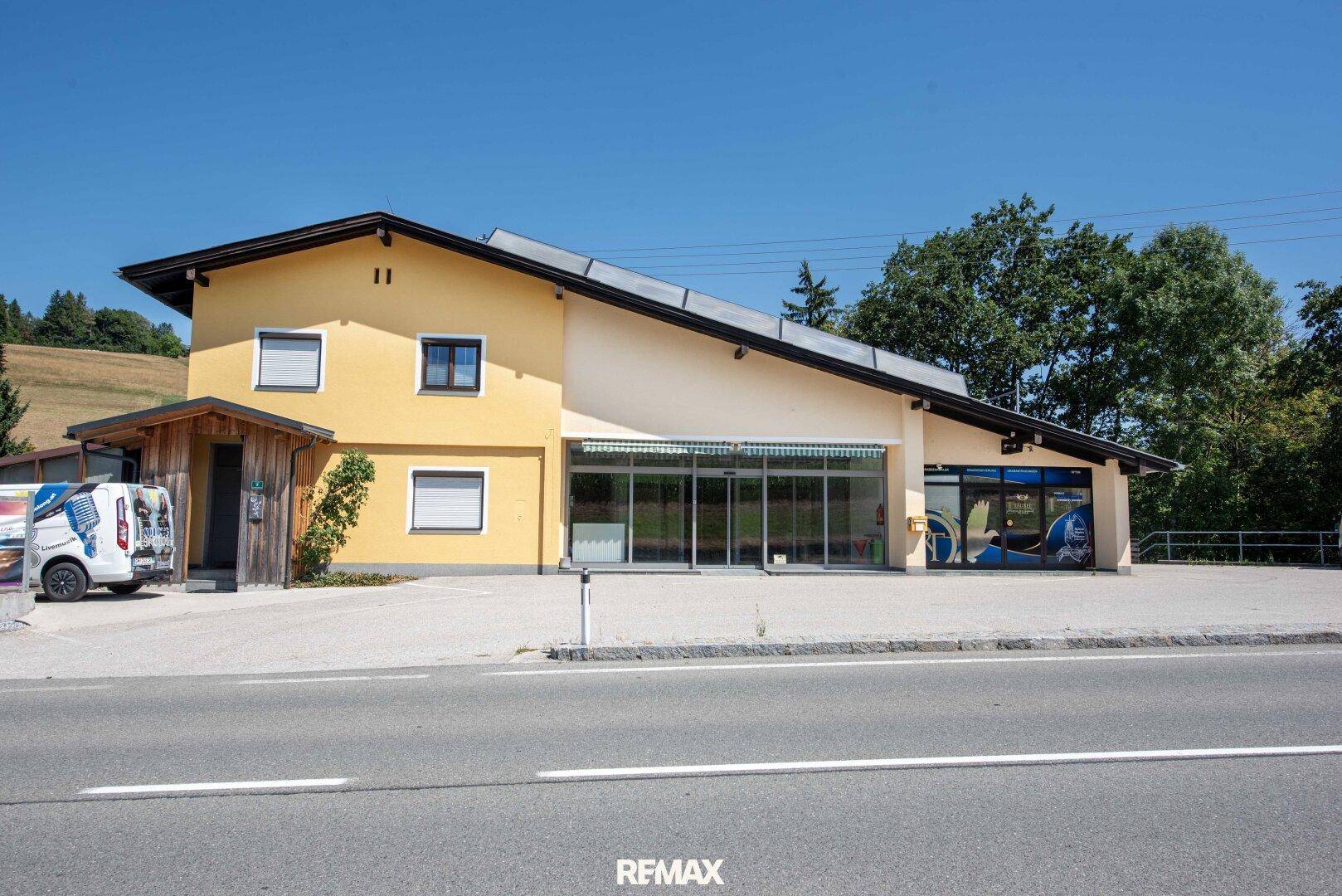 Gesamtansicht des gelben Gebäudes mit Solaranlage, Parkplätzen und angrenzender Straße vor grüner Hügellandschaft.
