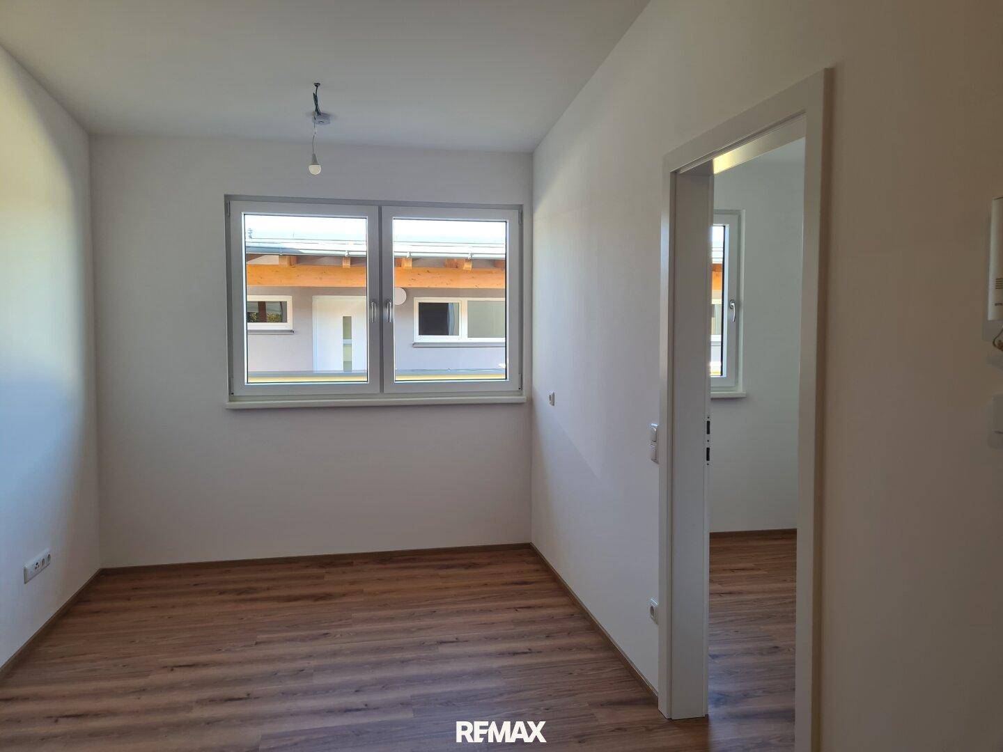 Leerer Raum mit Laminatboden und einem großen Fenster, das viel Tageslicht hereinlässt.