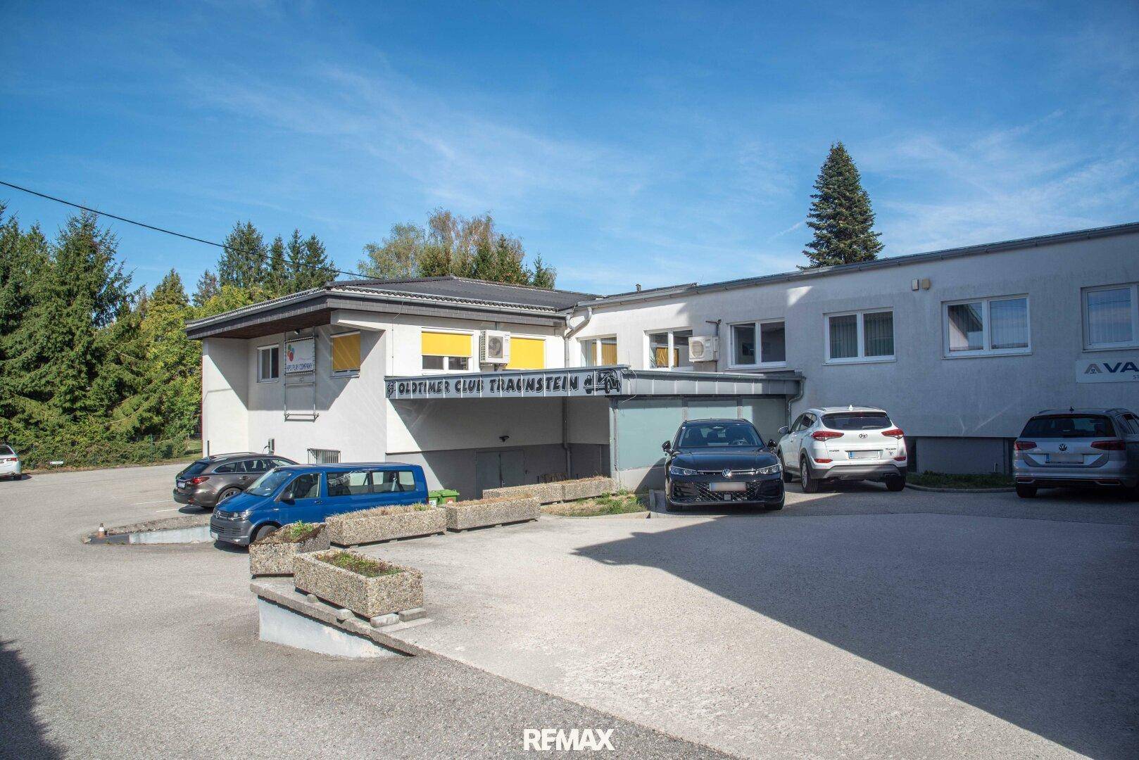 Rückansicht des Gebäudekomplexes mit weiteren Parkmöglichkeiten und umgebender Grünfläche unter blauem Himmel.