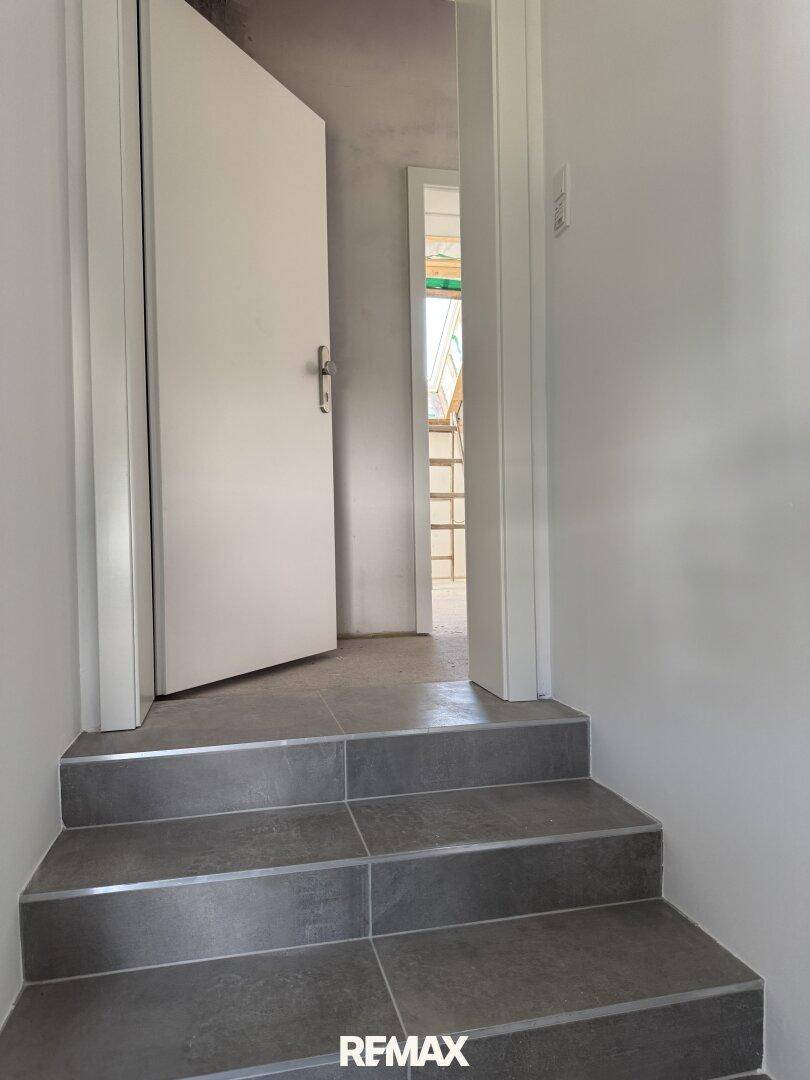Treppenaufgang mit grauen Fliesen und offener Tür zu einem im Bau befindlichen Dachgeschoss.