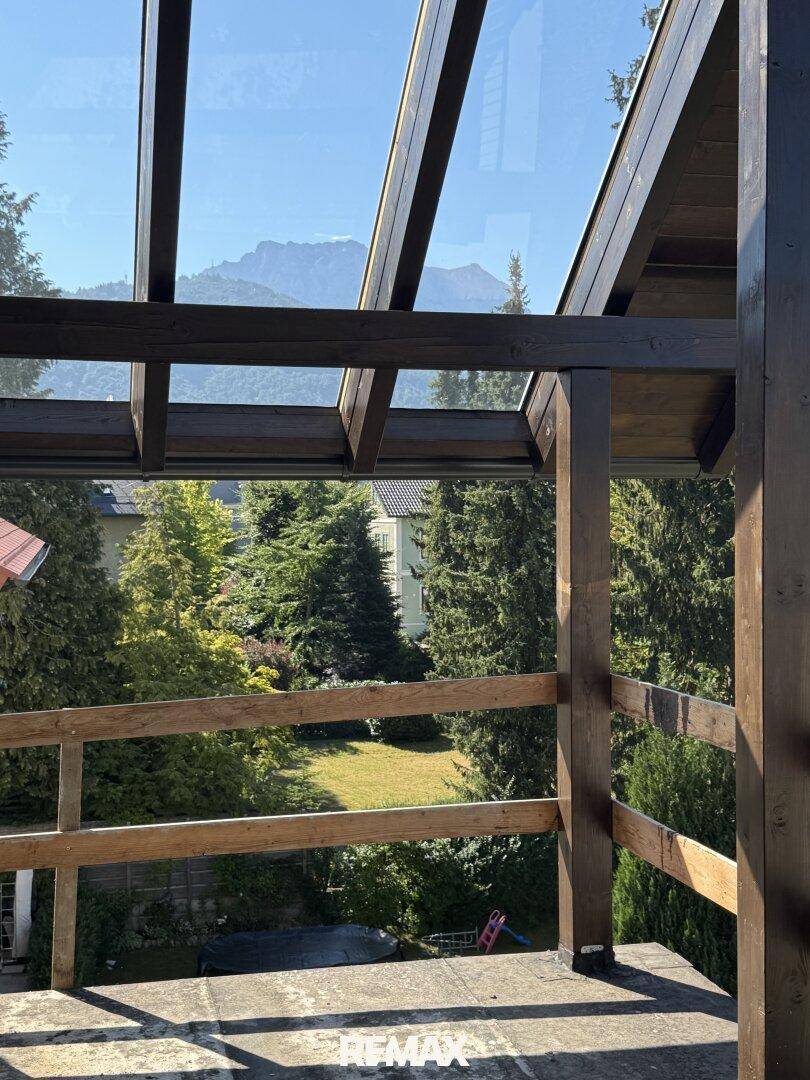Aussicht von einem Balkon mit Holzpergola und Glasdach auf grüne Bäume und ferne Berge.