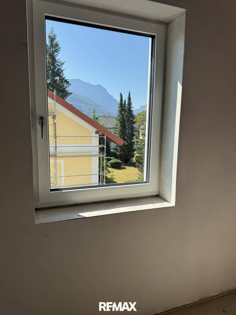 Innenraum mit Fensterblick auf ein gelbes Haus, grüne Bäume und eine beeindruckende Bergkulisse.