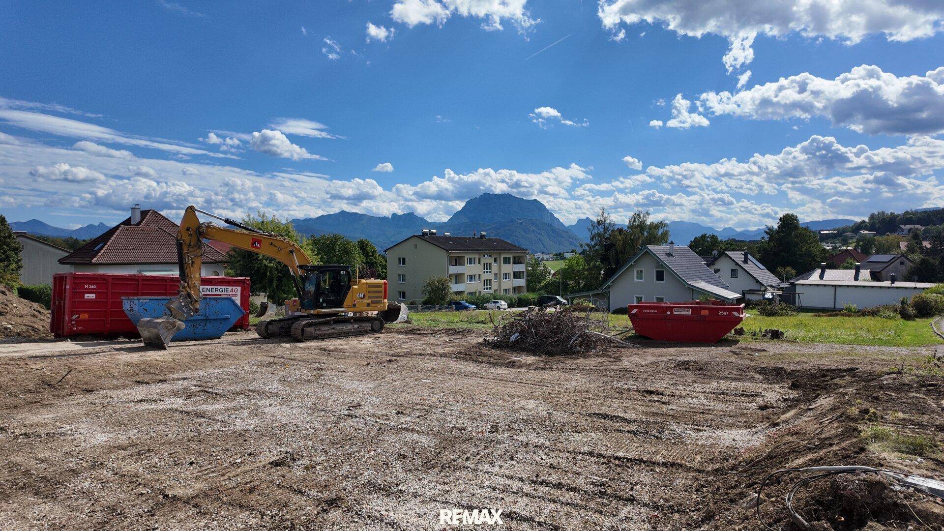 Baustelle mit Bagger und Containern, umgeben von Wohnhäusern und einer beeindruckenden Bergkulisse.