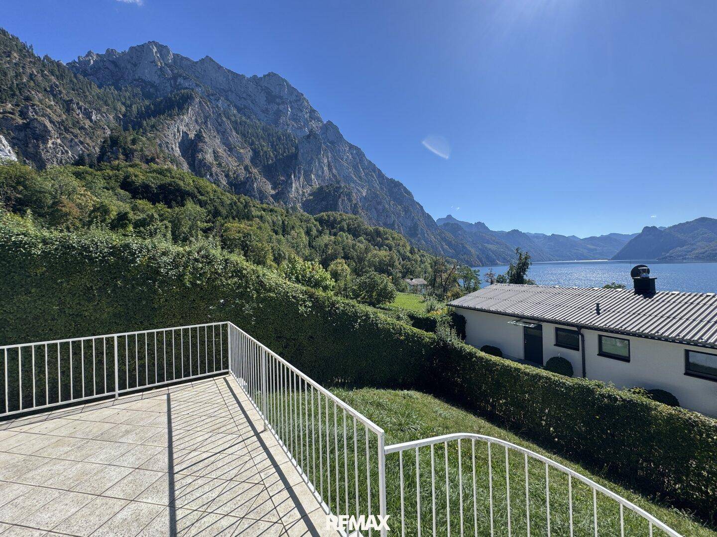 Sonniger Terrassenbereich mit Hecke und beeindruckendem Blick auf die majestätischen Berge und den See.