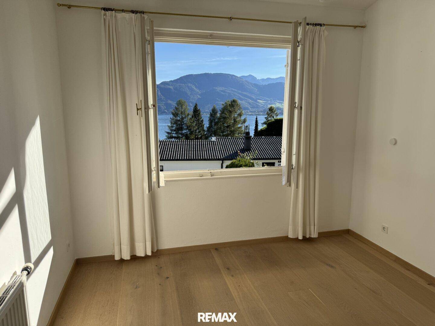 Schlafzimmer mit Holzboden, großem Fenster und malerischem Blick auf den See und die Berge.