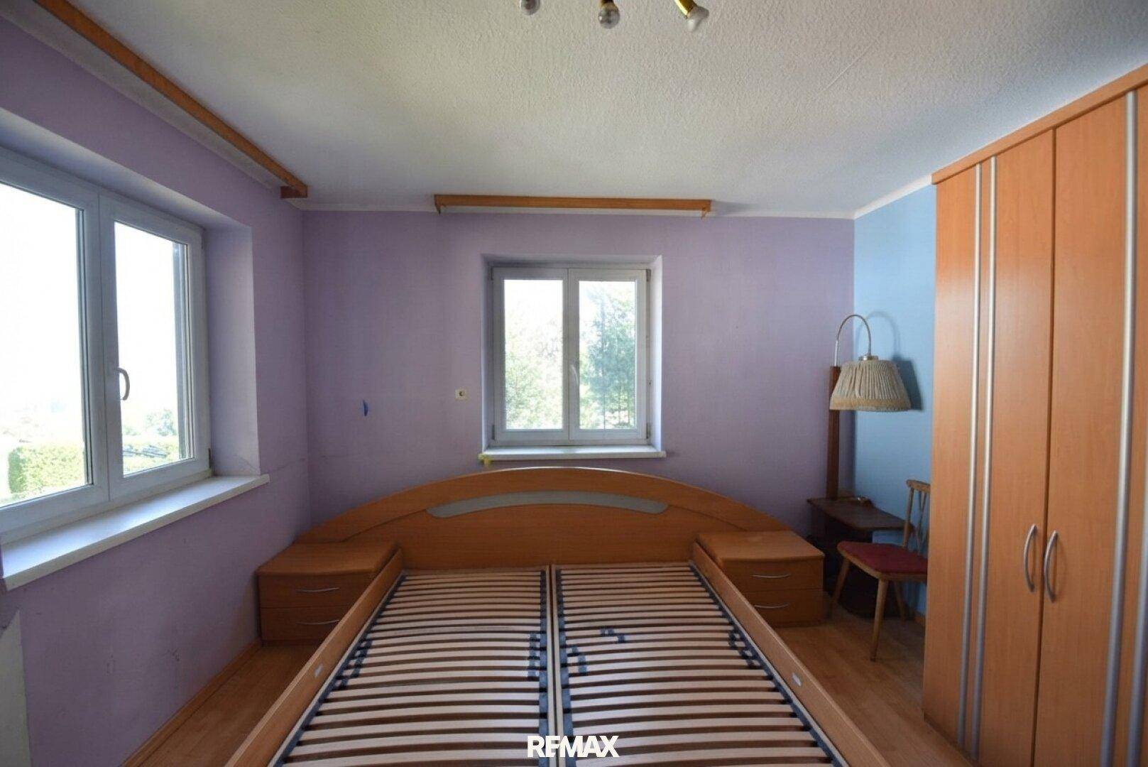 Schlafzimmer mit Doppelbett, Holzschrank und zwei Fenstern mit Blick ins Grüne.