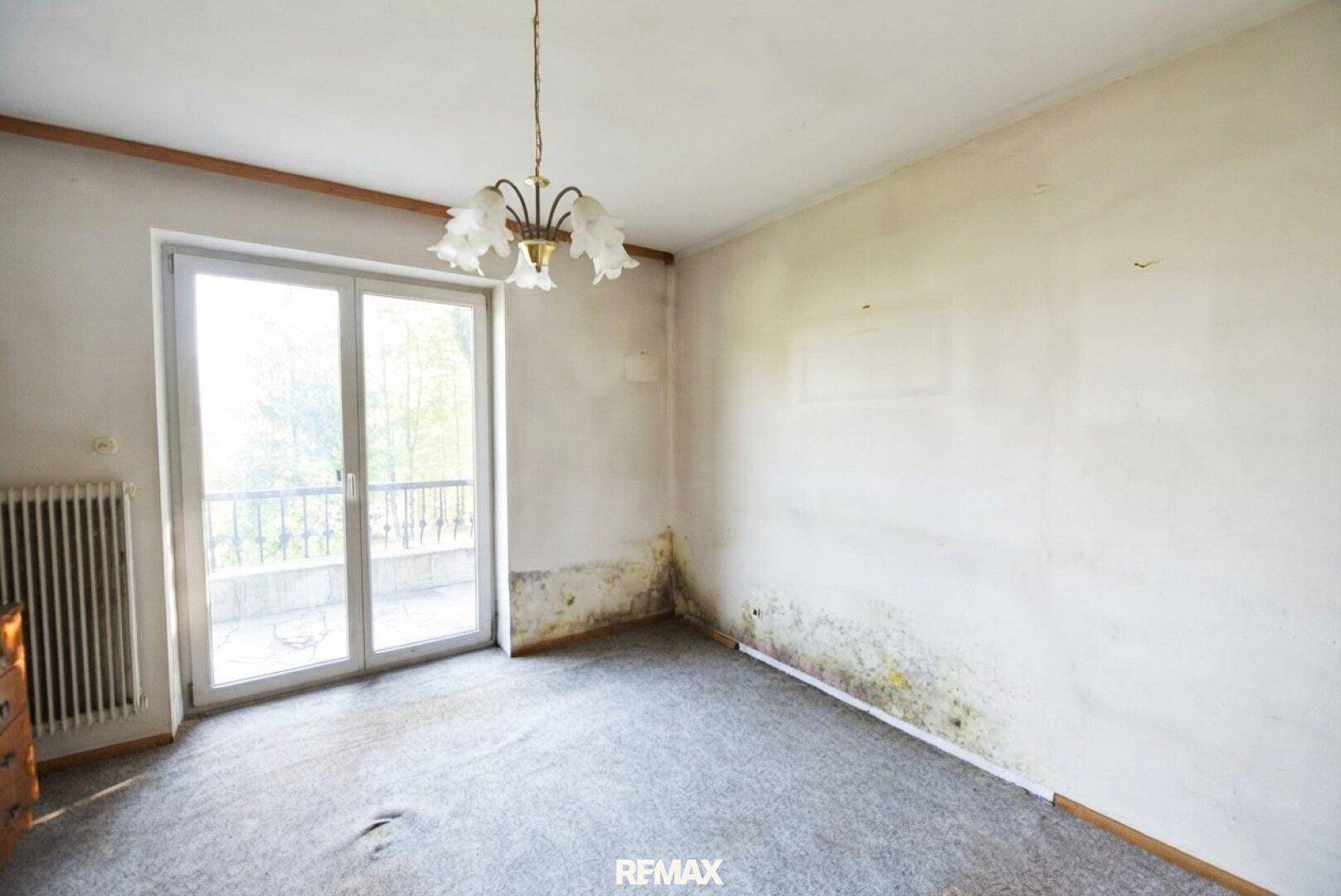 Zimmer mit Teppichboden, Balkontür und sichtbaren Feuchtigkeitsspuren an den Wänden.