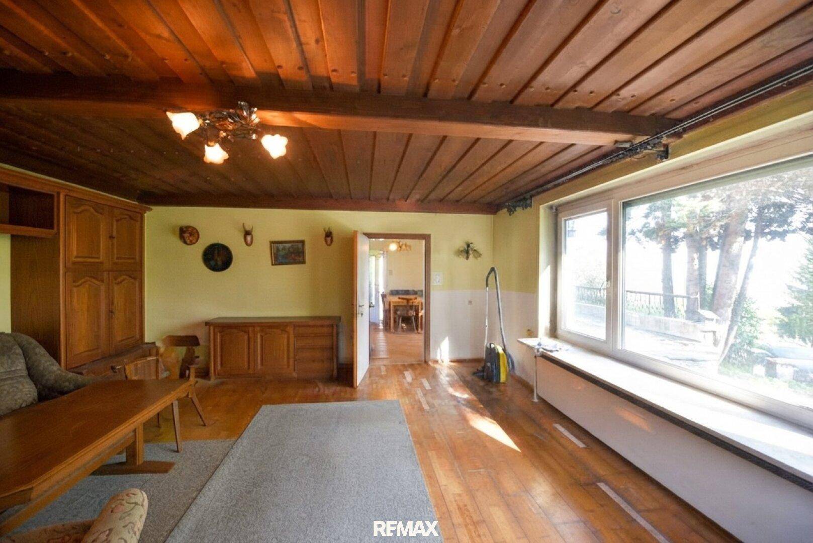 Gemütliches Wohnzimmer mit Holzbalkendecke, Parkettboden und großen Fenstern.