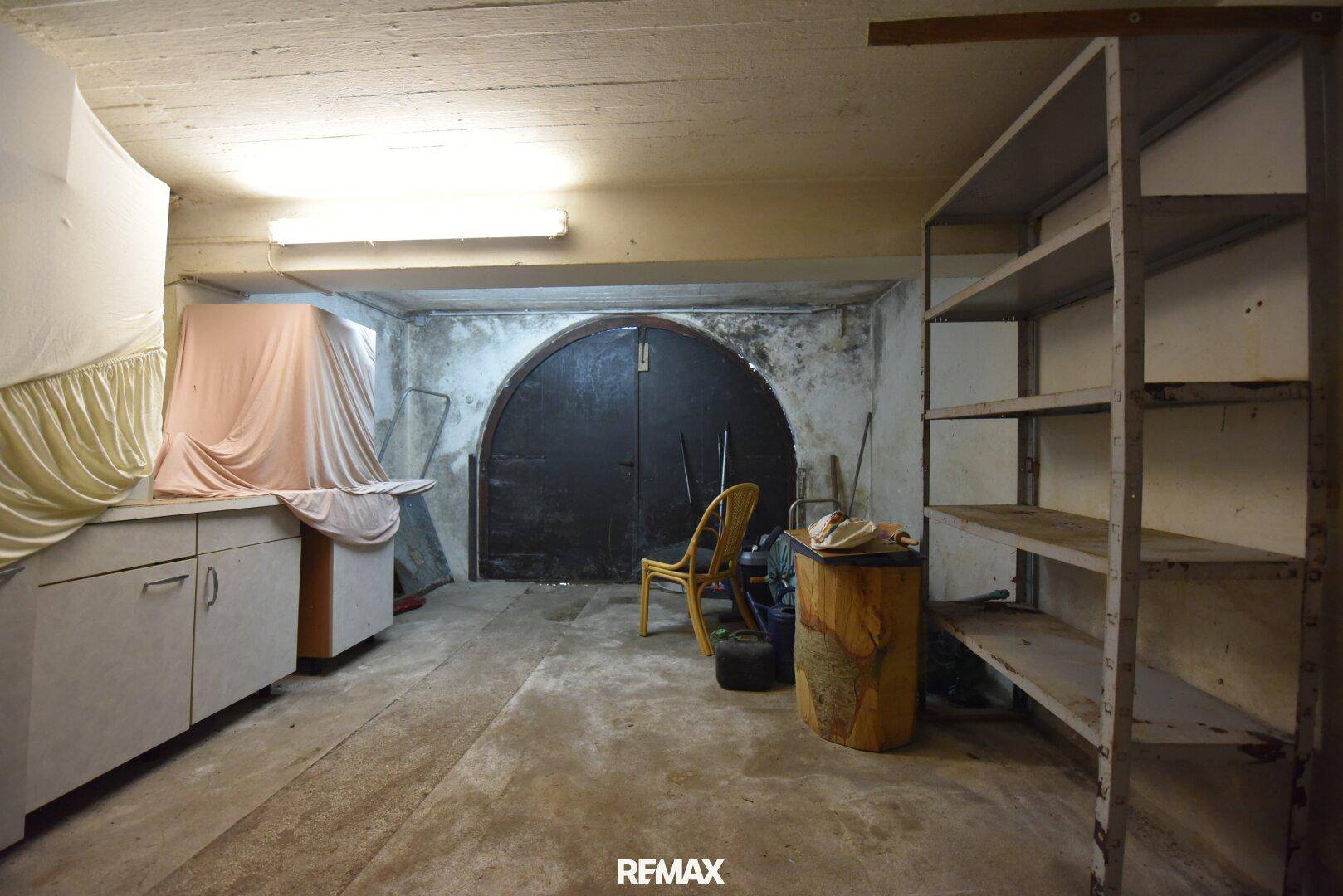 Kellerraum mit Betonboden, Regalen und einem großen, gewölbten Tor.