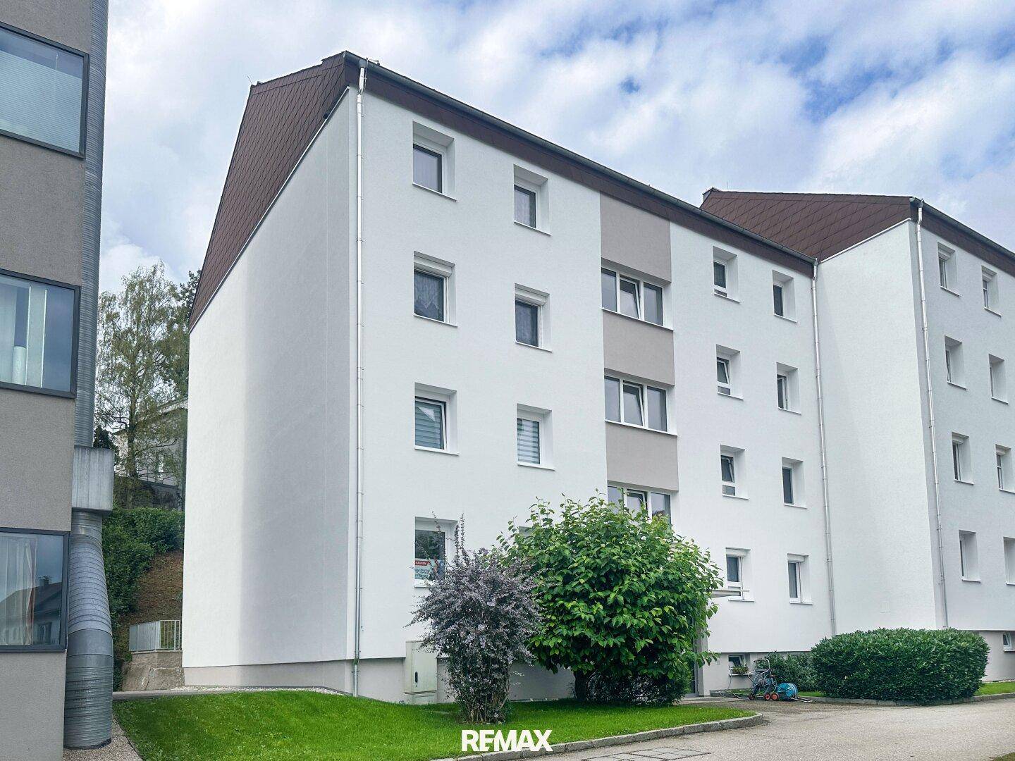 Mehrfamilienhaus mit weißer Fassade, vielen Fenstern und einem Satteldach in grüner Umgebung.