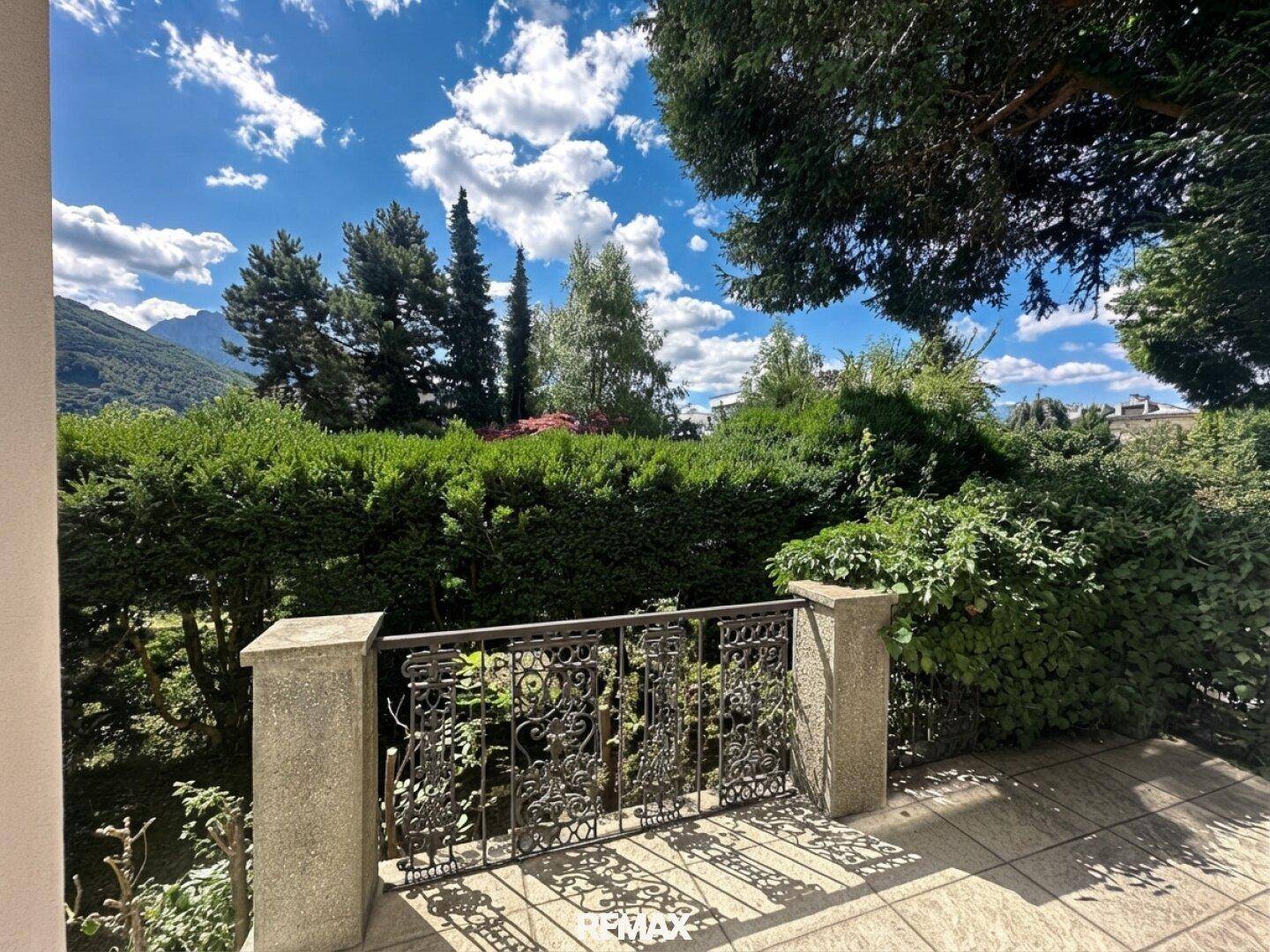 Gepflasterte Terrasse mit schmiedeeisernem Geländer, umgeben von grünen Hecken und Bergblick.