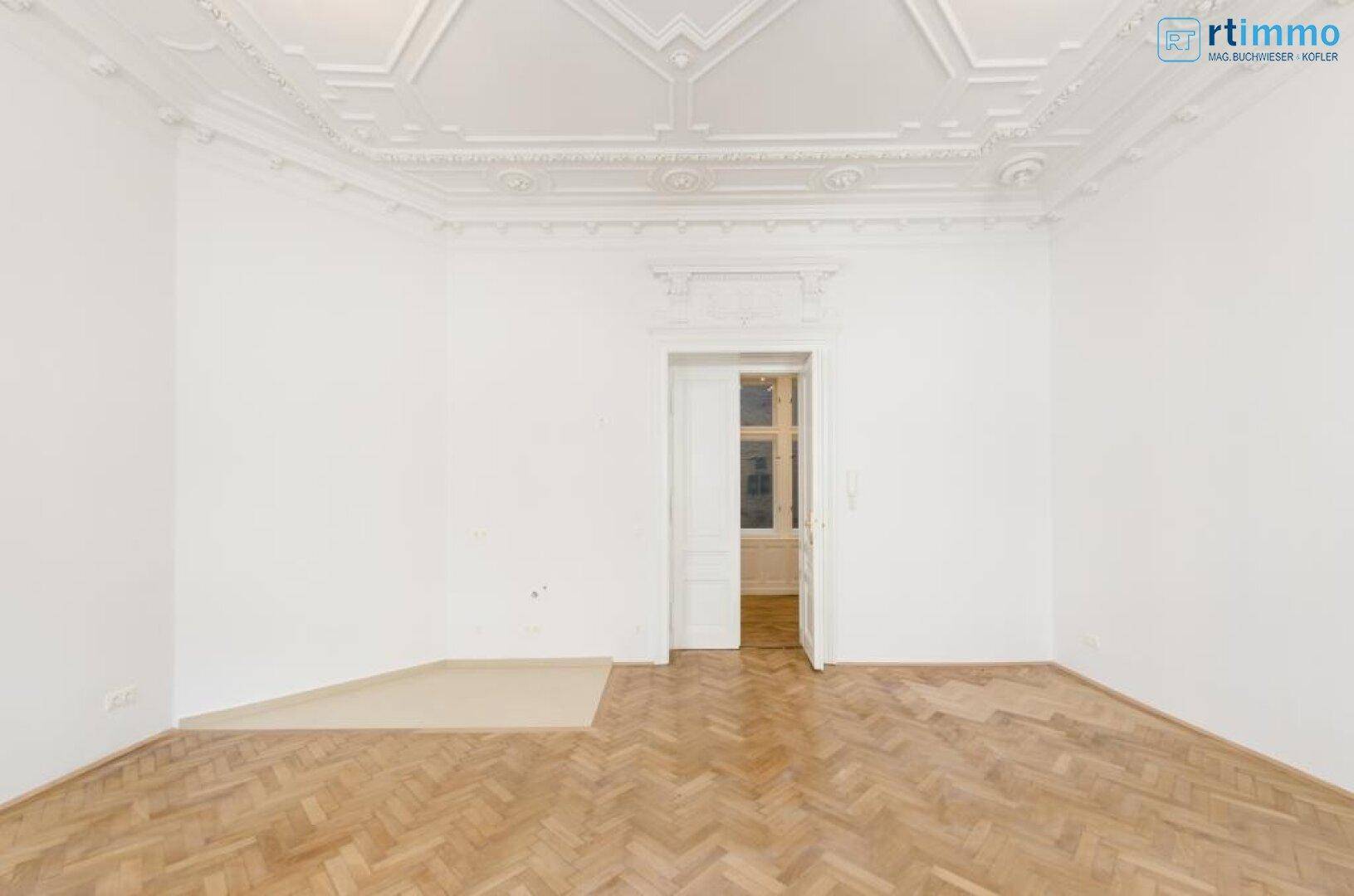 Geräumiger Altbauraum mit Fischgrätparkett, weiß gestrichenen Wänden und einer eleganten Stuckdecke.