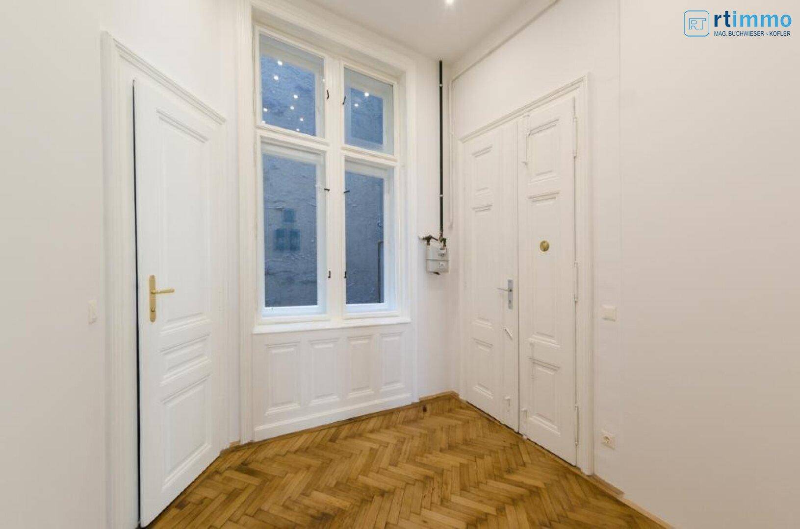 Kleiner Flur mit Fischgrätparkett, einem Fenster und zwei weißen Türen in einem Altbau.