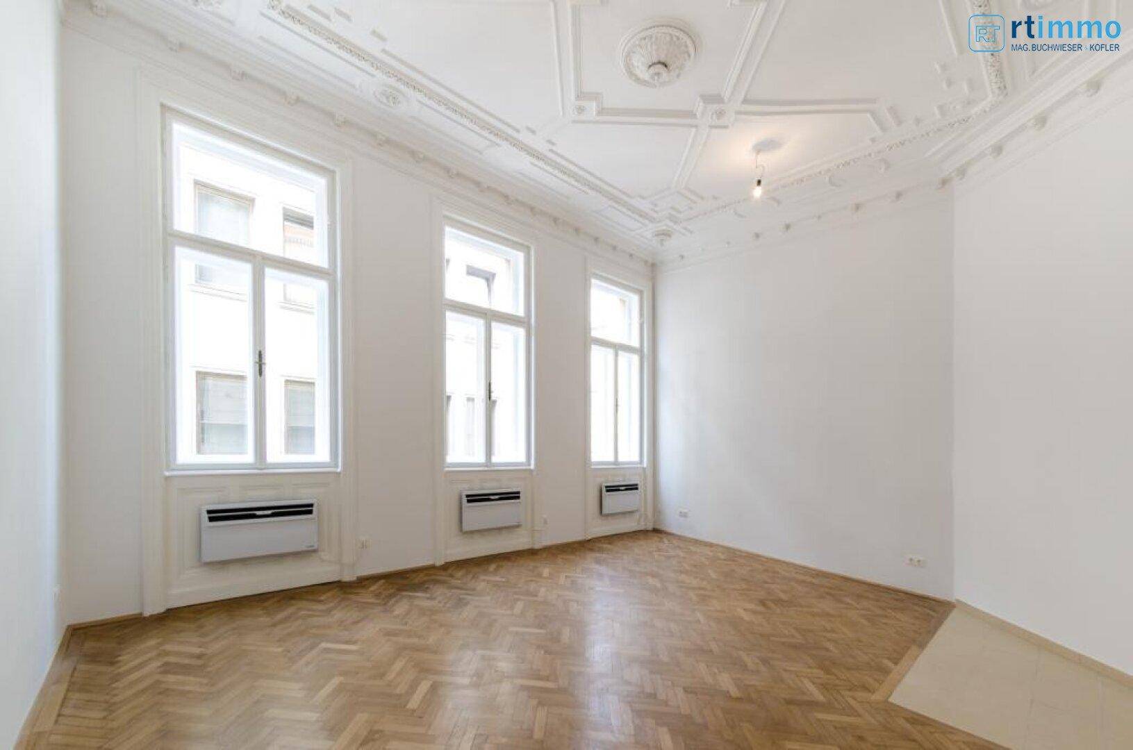 Geräumiger leerer Raum mit Fischgrätparkett, hohen Fenstern und einer kunstvollen Stuckdecke.