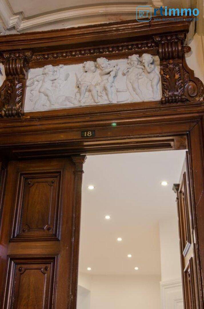 Prunkvoller Holztürrahmen mit figürlichem Relief, der in einen modern beleuchteten Raum führt.