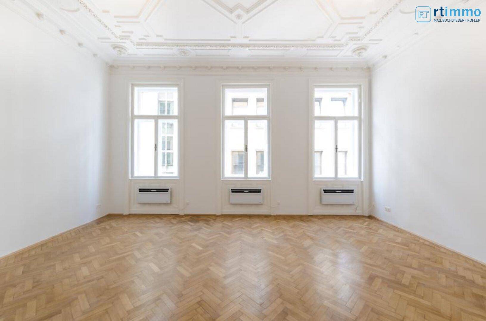 Heller, leerer Raum mit Fischgrätparkett, hohen Fenstern und einer reich verzierten Stuckdecke.