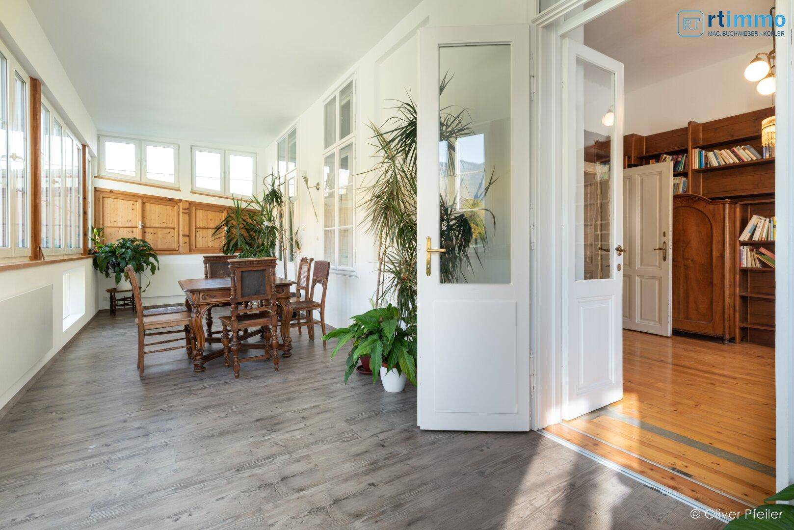 Geräumiger Wintergarten mit Esstisch und Zugang zu einem weiteren Zimmer mit Holzmöbeln.