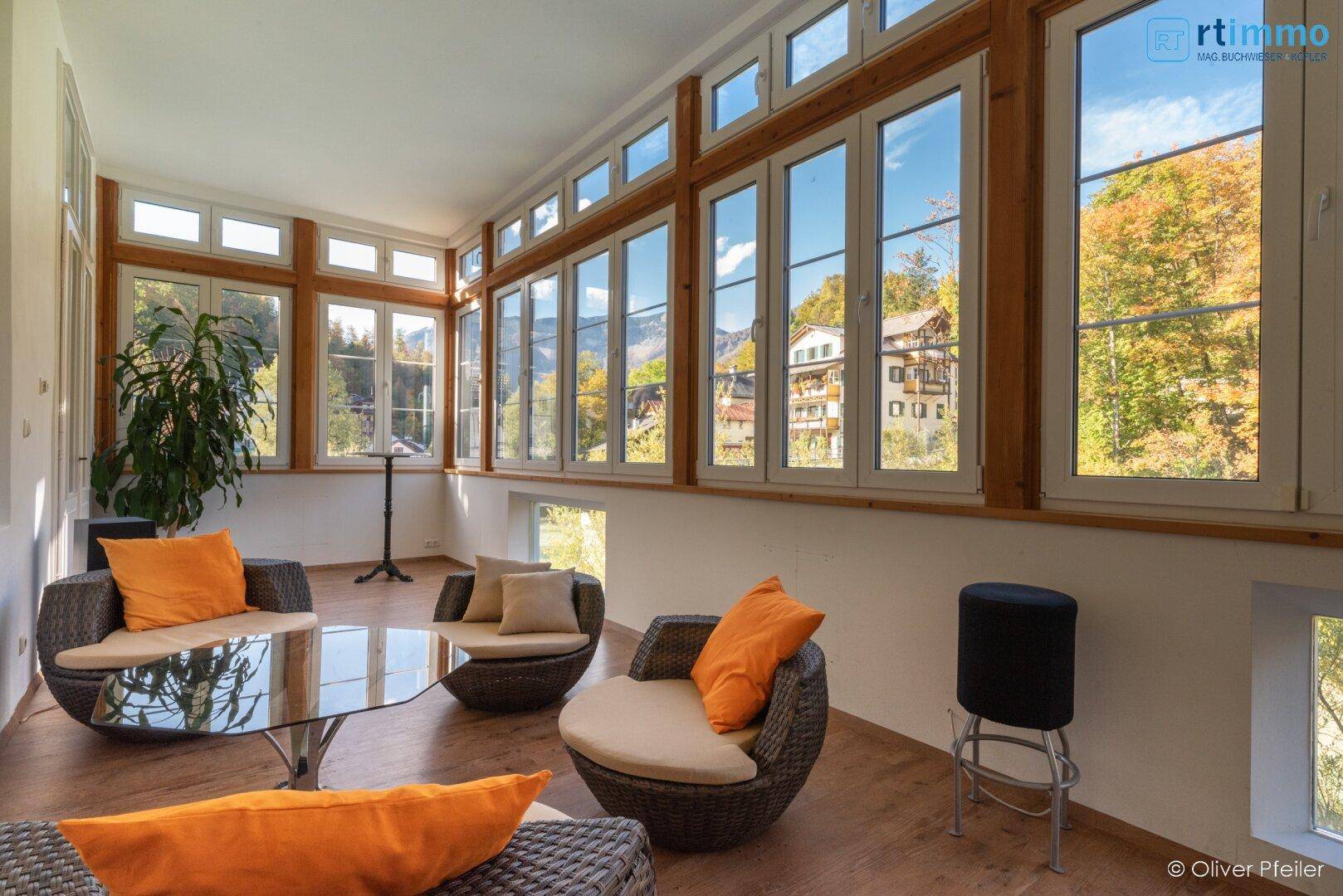 Heller Wintergarten mit Rattanmöbeln, Holzboden und Panoramablick auf die Natur.