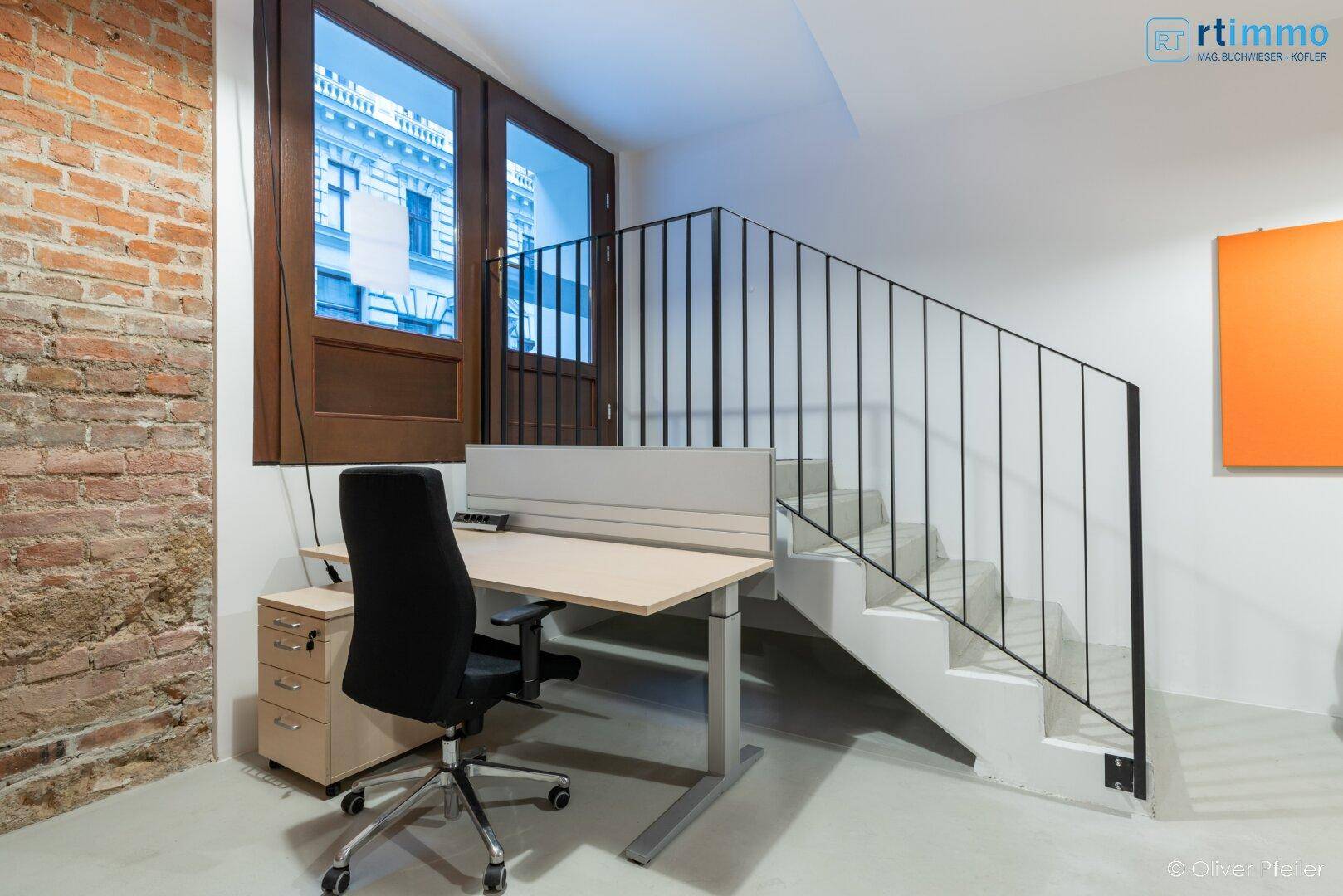Arbeitsbereich mit Schreibtisch, Stuhl und einer Treppe, die zu einem oberen Bereich führt, mit großen Fenstern.