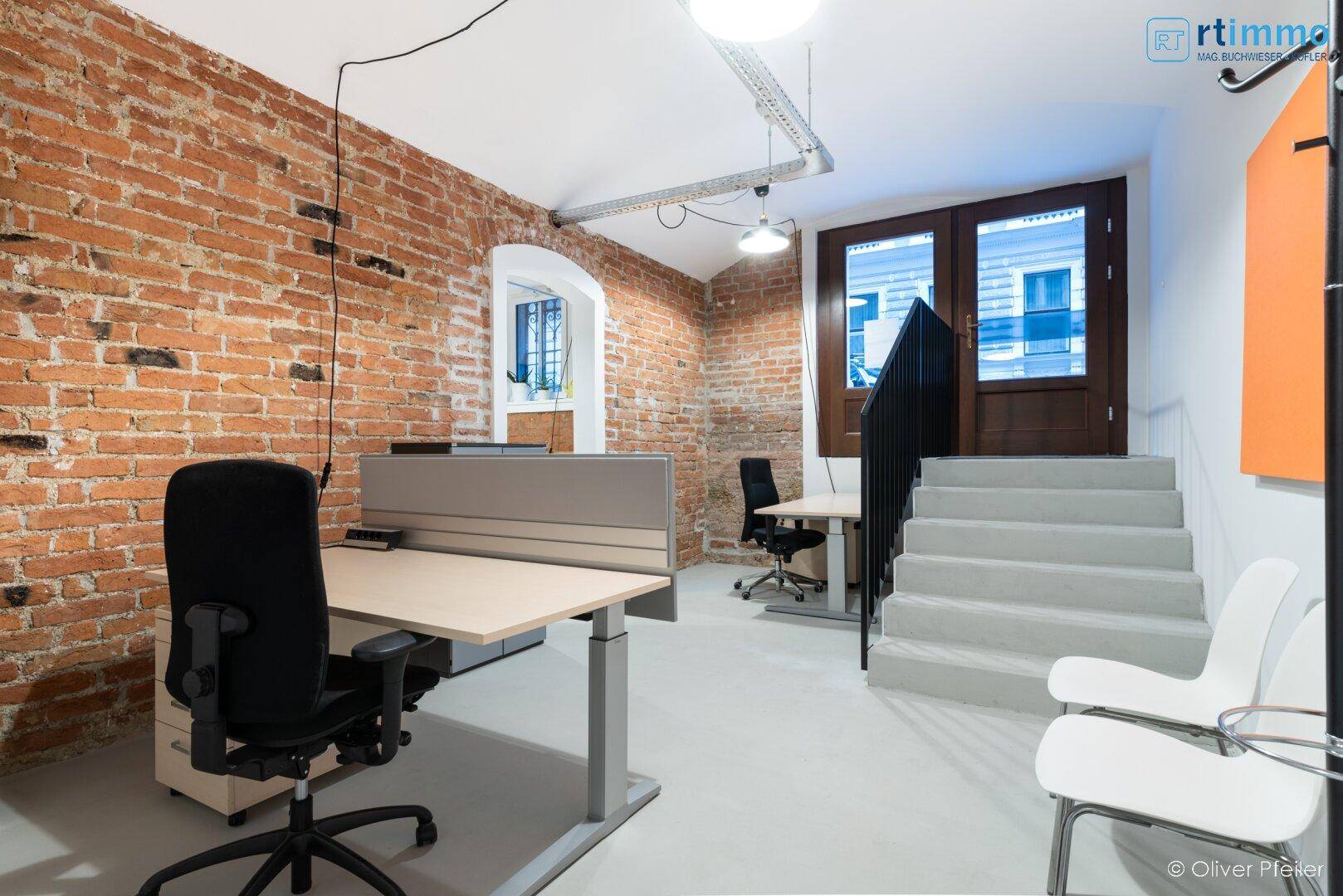 Offener Bürobereich mit freiliegenden Ziegelwänden, mehreren Arbeitsplätzen und einer modernen Treppe.