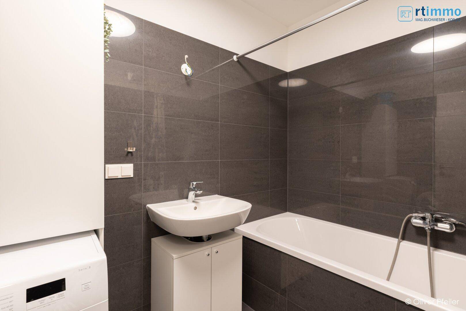 Badezimmer mit dunklen Fliesen, Badewanne und Waschbecken, inklusive Waschmaschine.